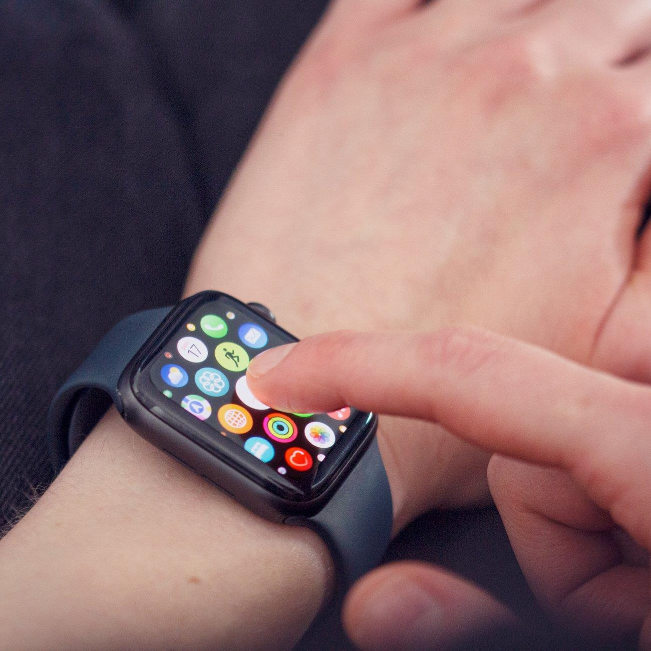Ein Finger berührt den Bildschirm einer Smartwatch, die mehrere App-Symbole auf einer dunklen Uhr mit blauem Armband zeigt.