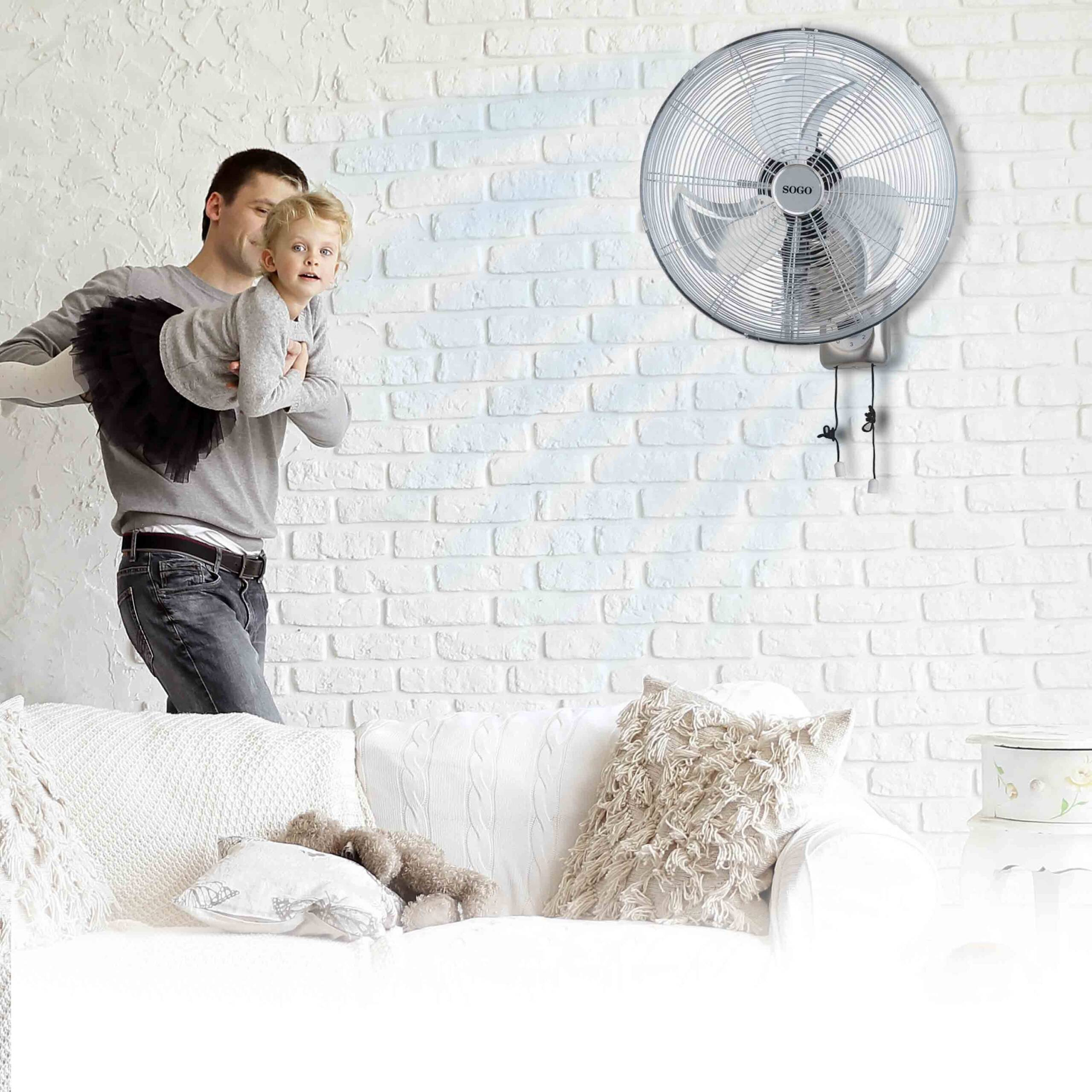 Hombre sosteniendo a una niña cerca de un ventilador de pared sobre una pared de ladrillos blancos.