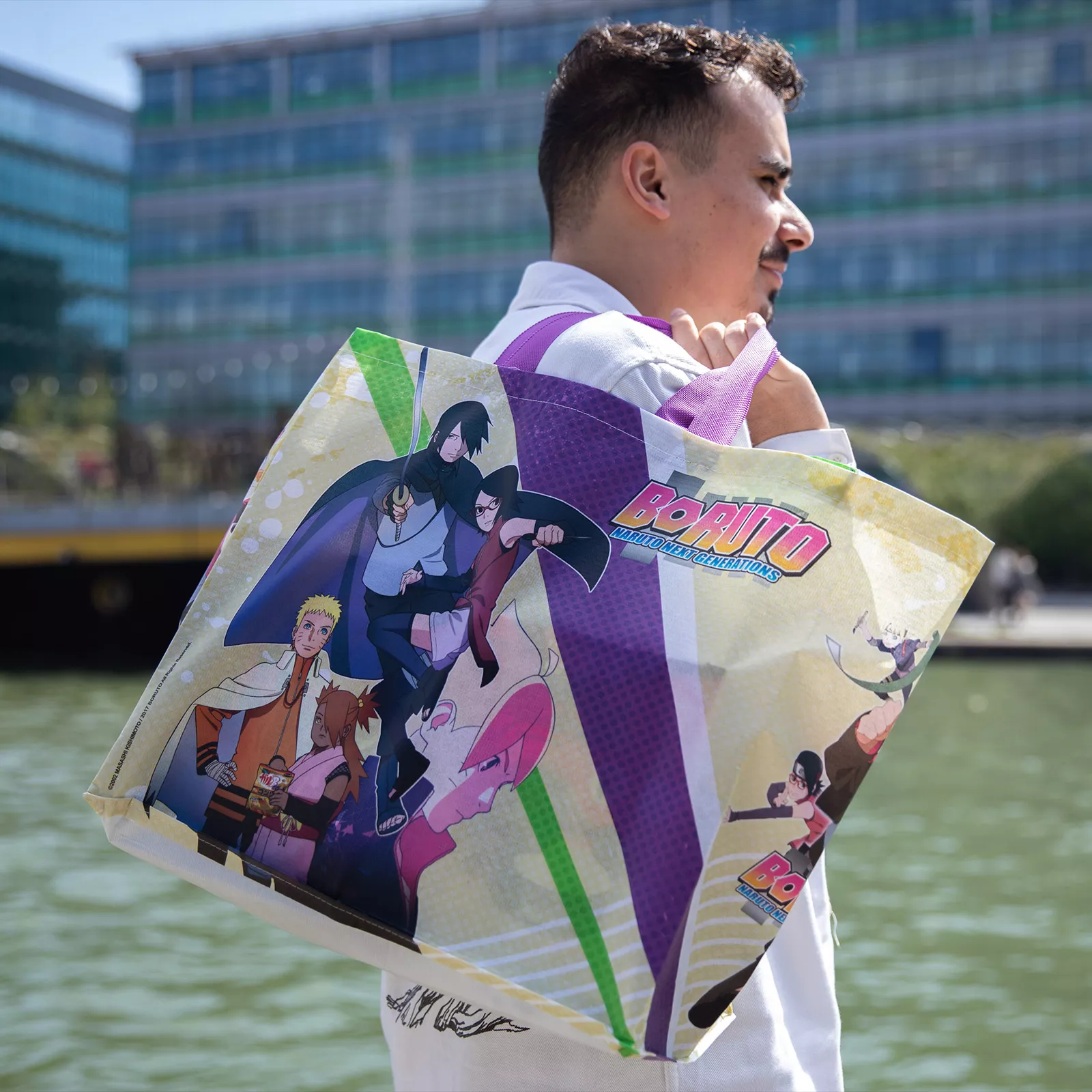 Person hält eine Boruto Tragetasche mit Anime-Charakteren. Der Hintergrund ist eine Stadt und ein Fluss.