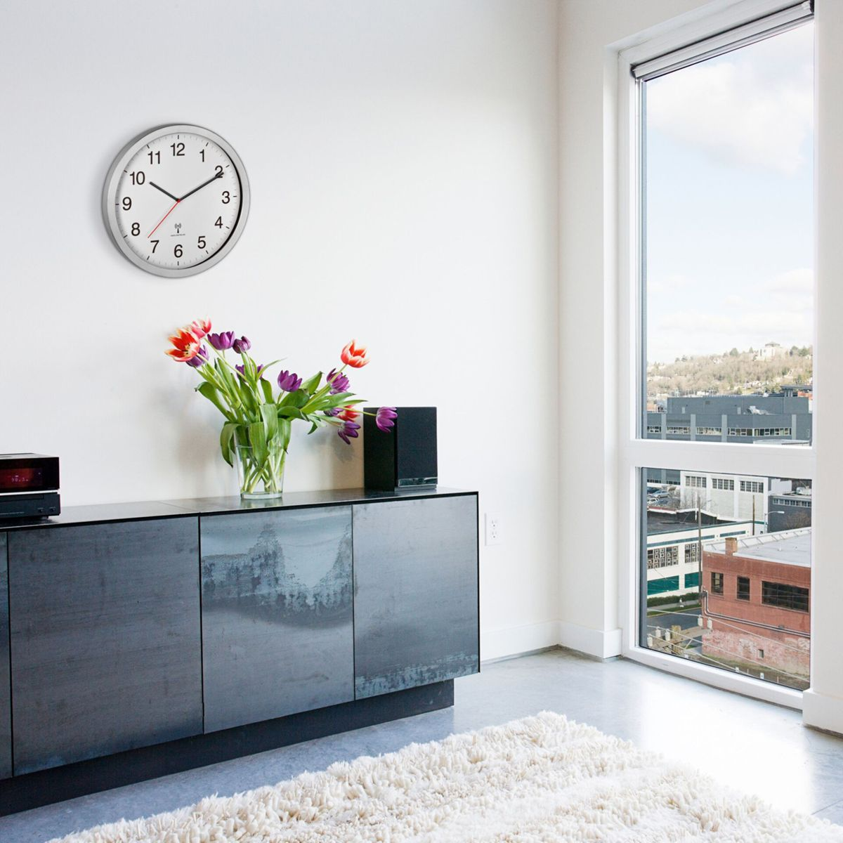 Ein Raum mit einer Uhr, Blumen in einer Vase und einem schwarzen Schrank. Rechts ist ein Fenster mit Aussicht.