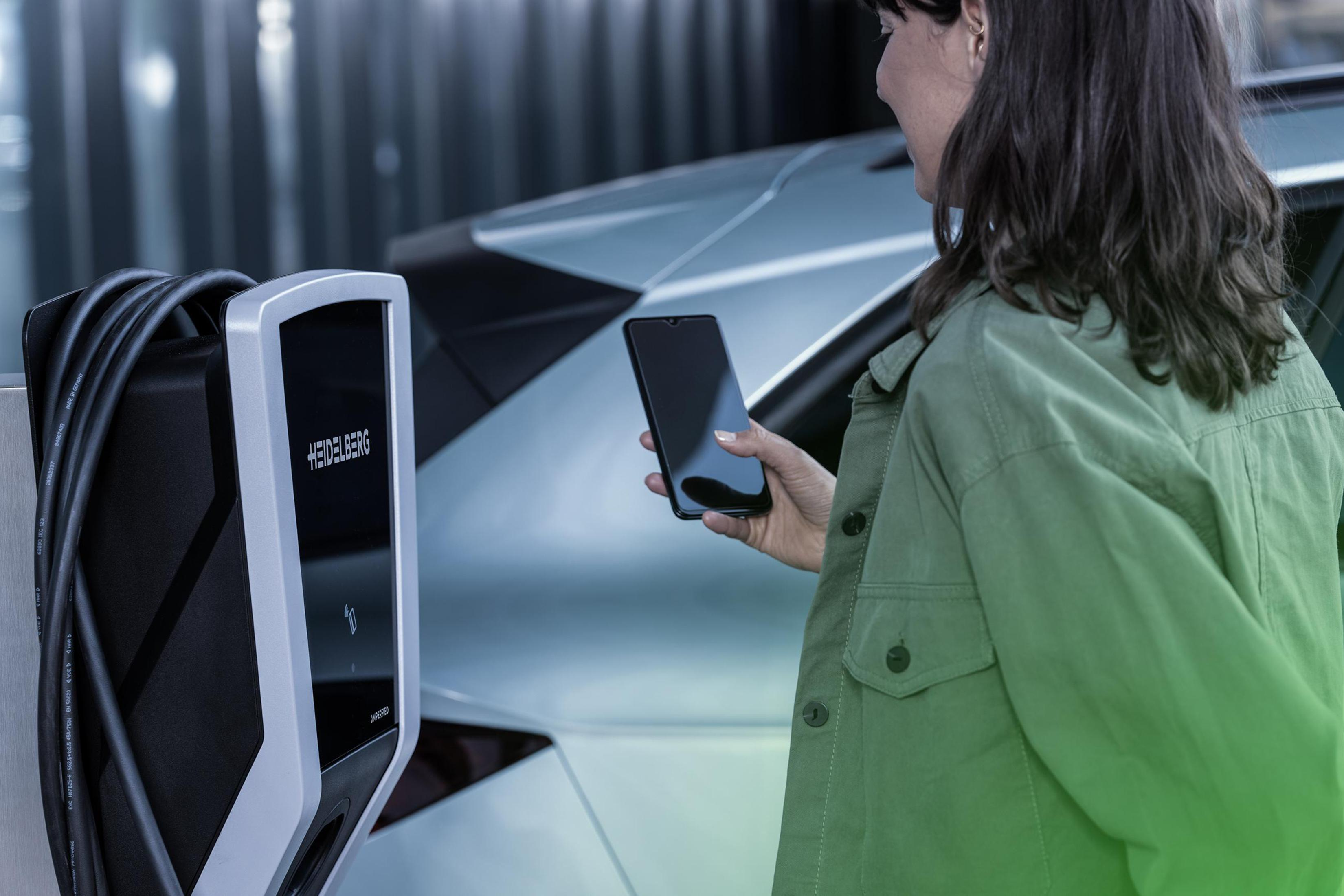 Eine Frau hält ein Telefon in der Nähe eines Heidelberg-Elektrofahrzeugladegeräts, neben einem silbernen Auto in einem Showroom.