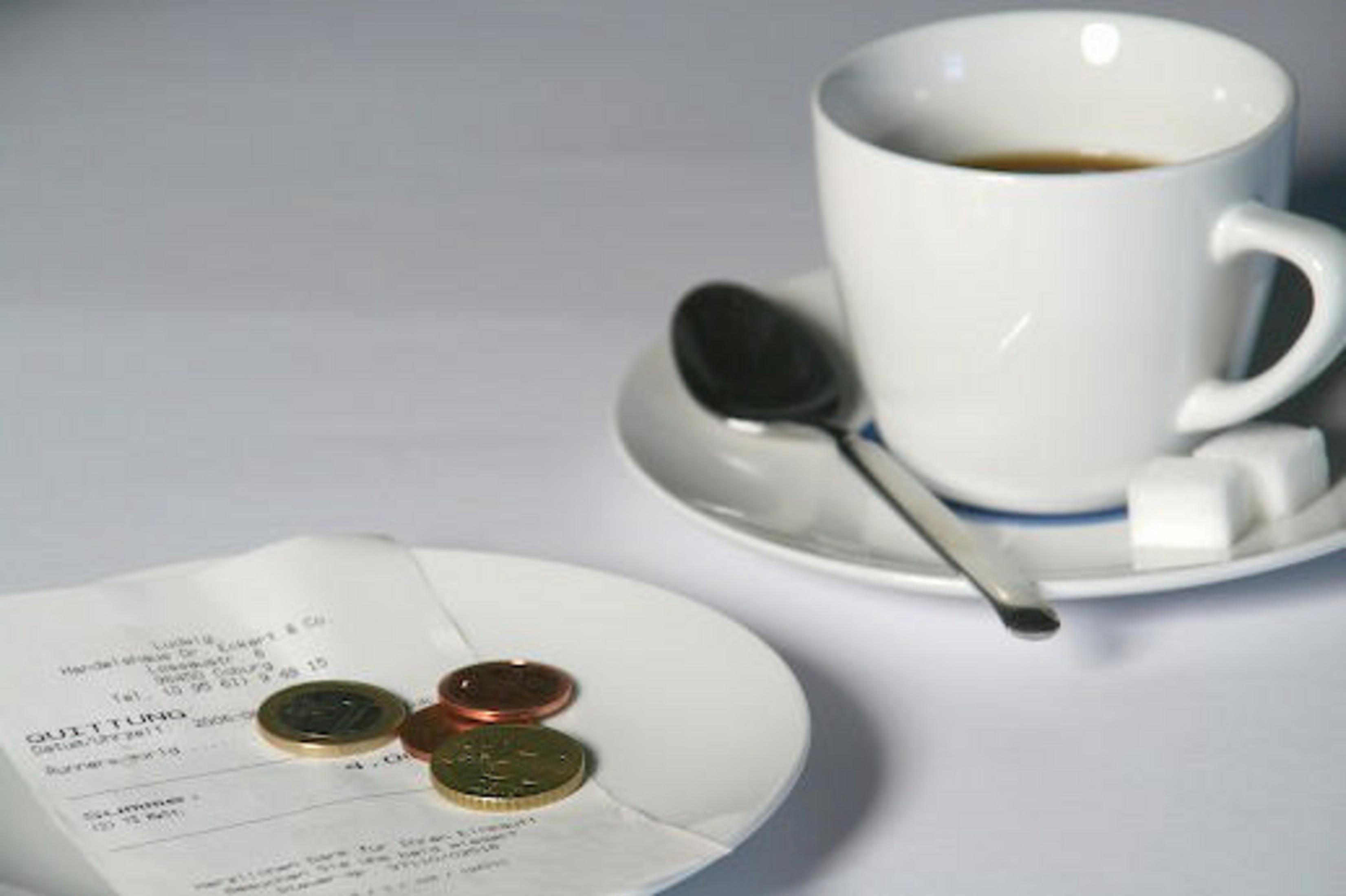Eine weiße Kaffeetasse und Untertasse mit einem schwarzen Löffel. Daneben ein Beleg mit Münzen auf einem Teller.