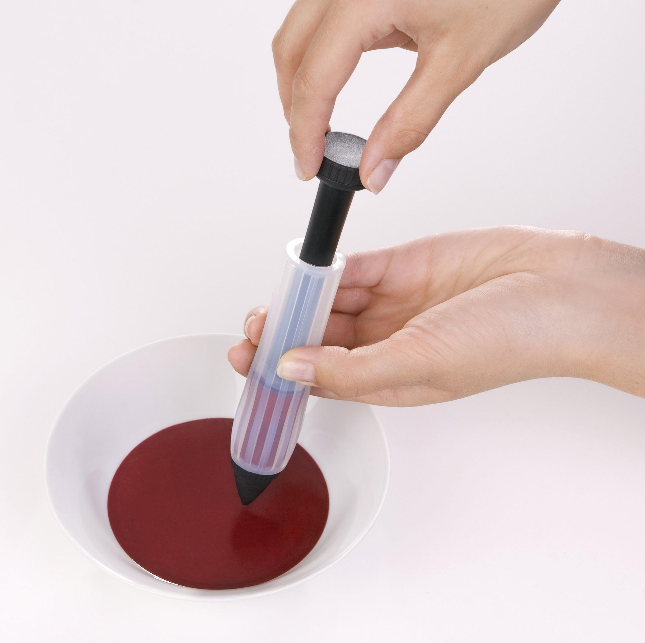 Eine Hand füllt einen Dekorationsstift mit roter Flüssigkeit. Der Stift liegt in einer weißen Schüssel.