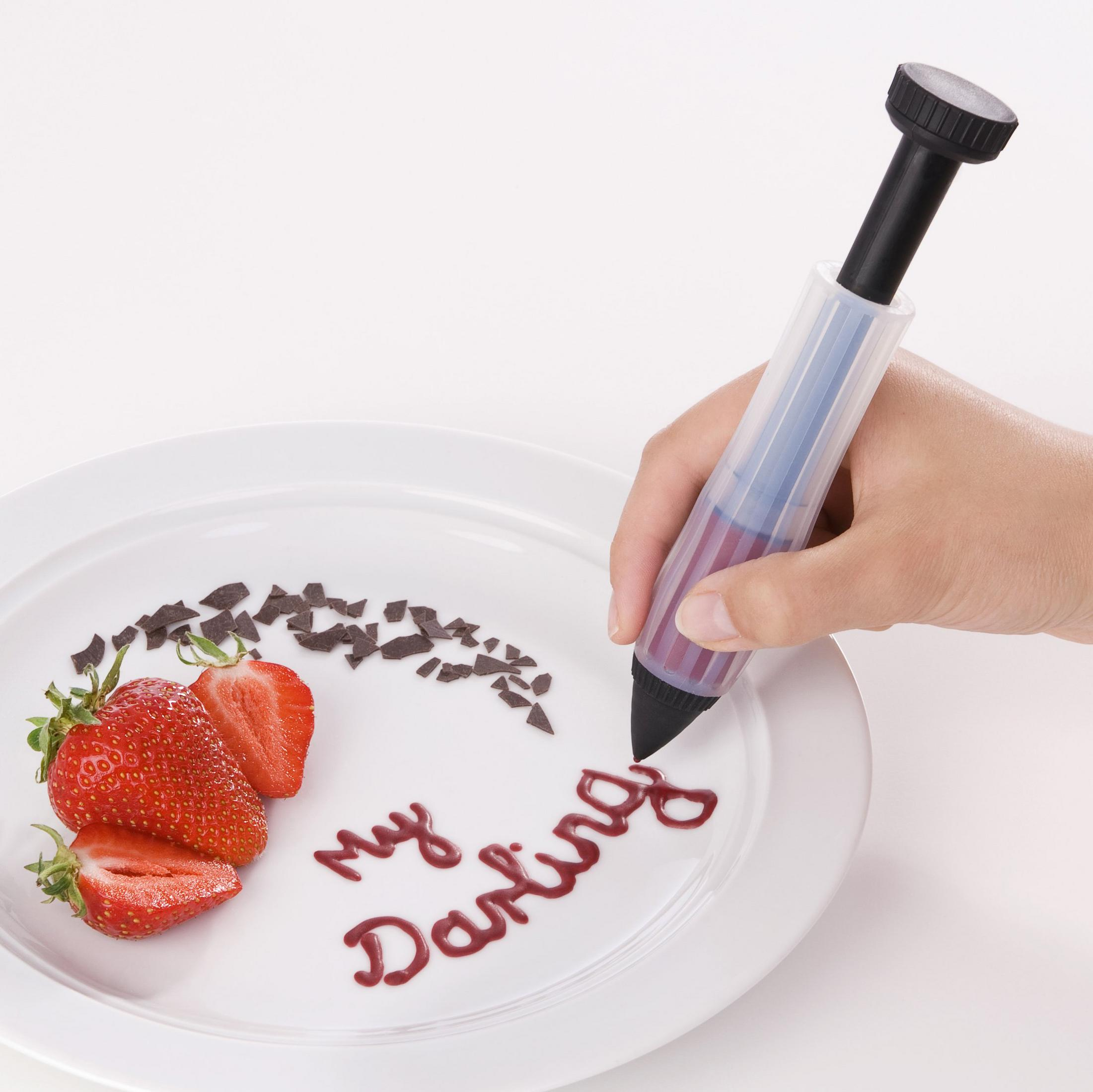 Eine Hand schreibt "My Darling" mit rotem Zuckerguss auf einen Teller. Erdbeeren und Schokostücke sind vorhanden.