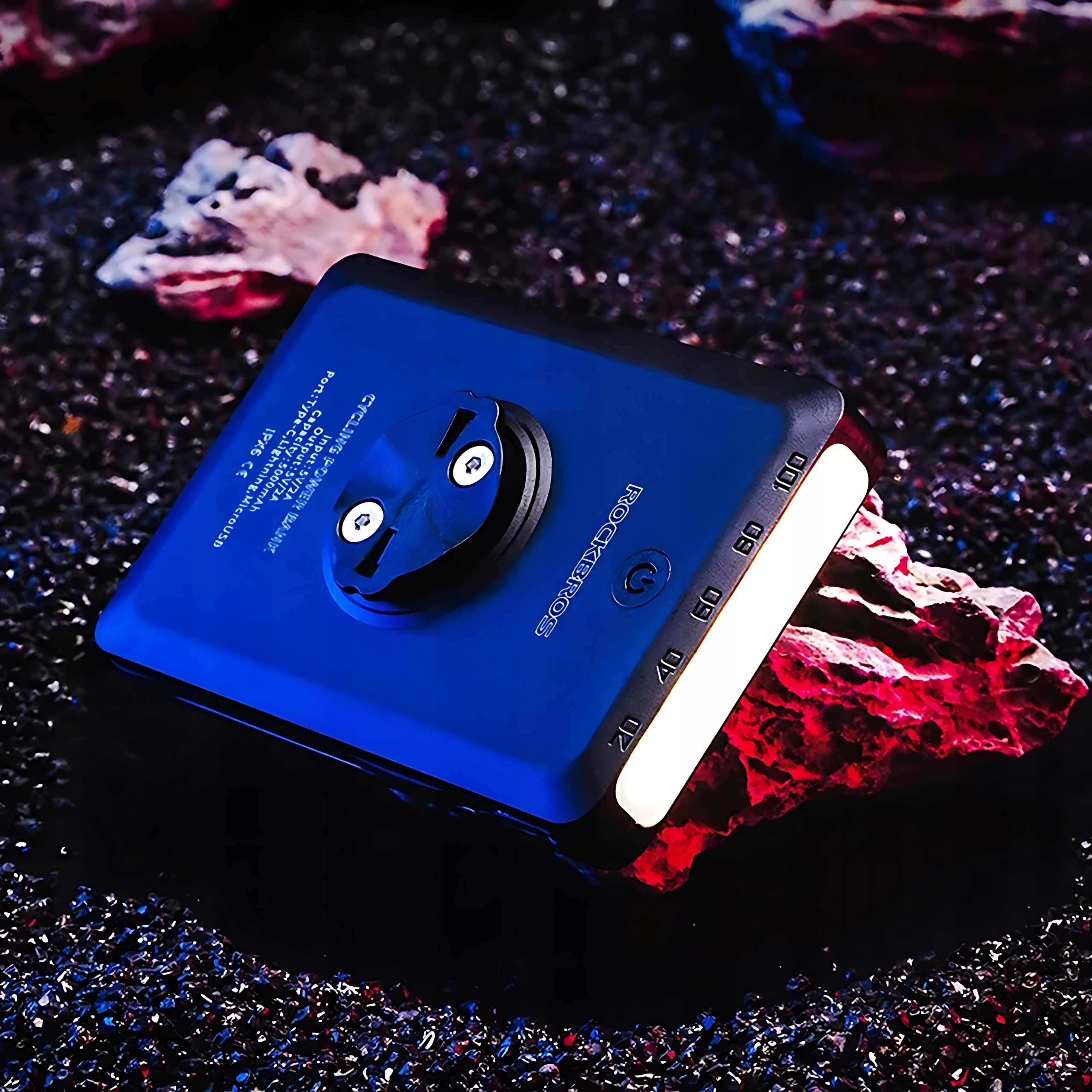 Niebieskie urządzenie z logo Rockbros na skalistej powierzchni. Urządzenie ma biały pasek świetlny i przycisk zasilania.