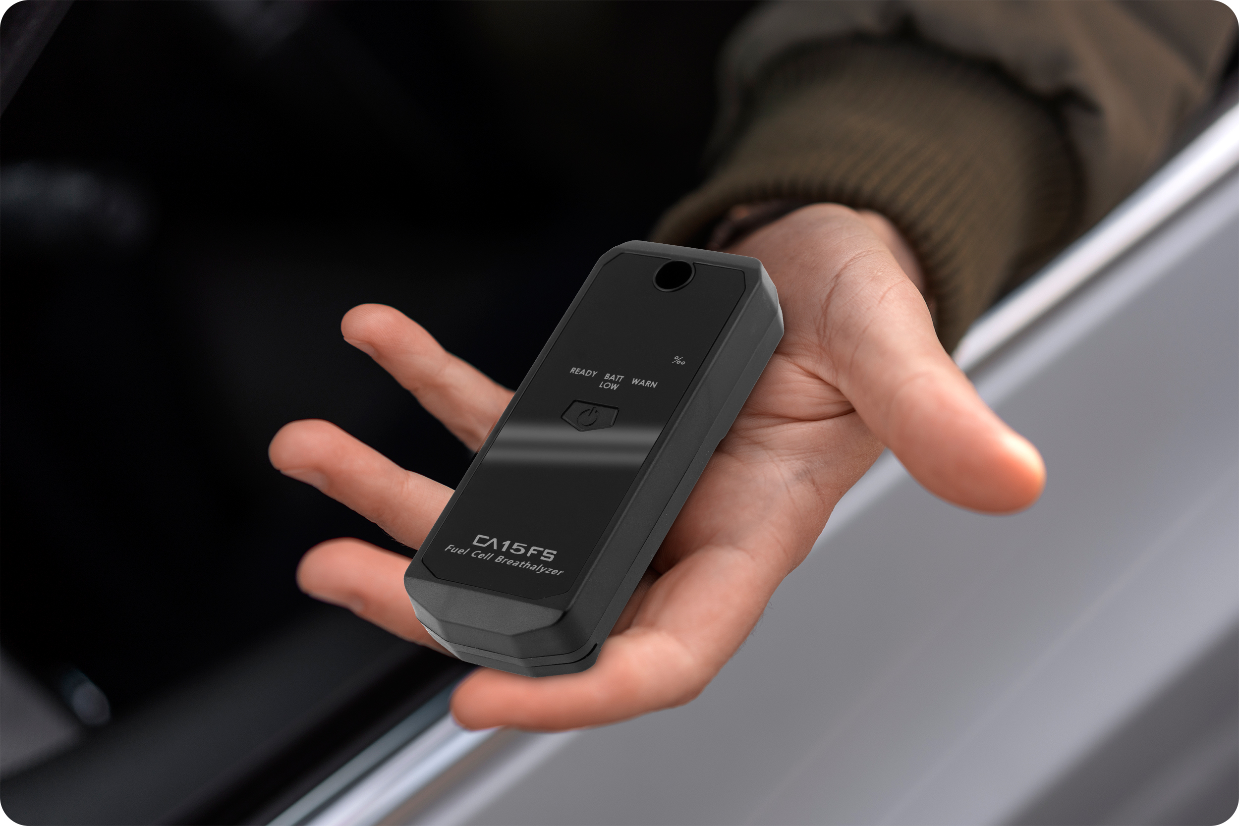 Ręka trzymająca czarny alkomat obok szyby samochodu. Alkomat oznaczony jest CA15FS.