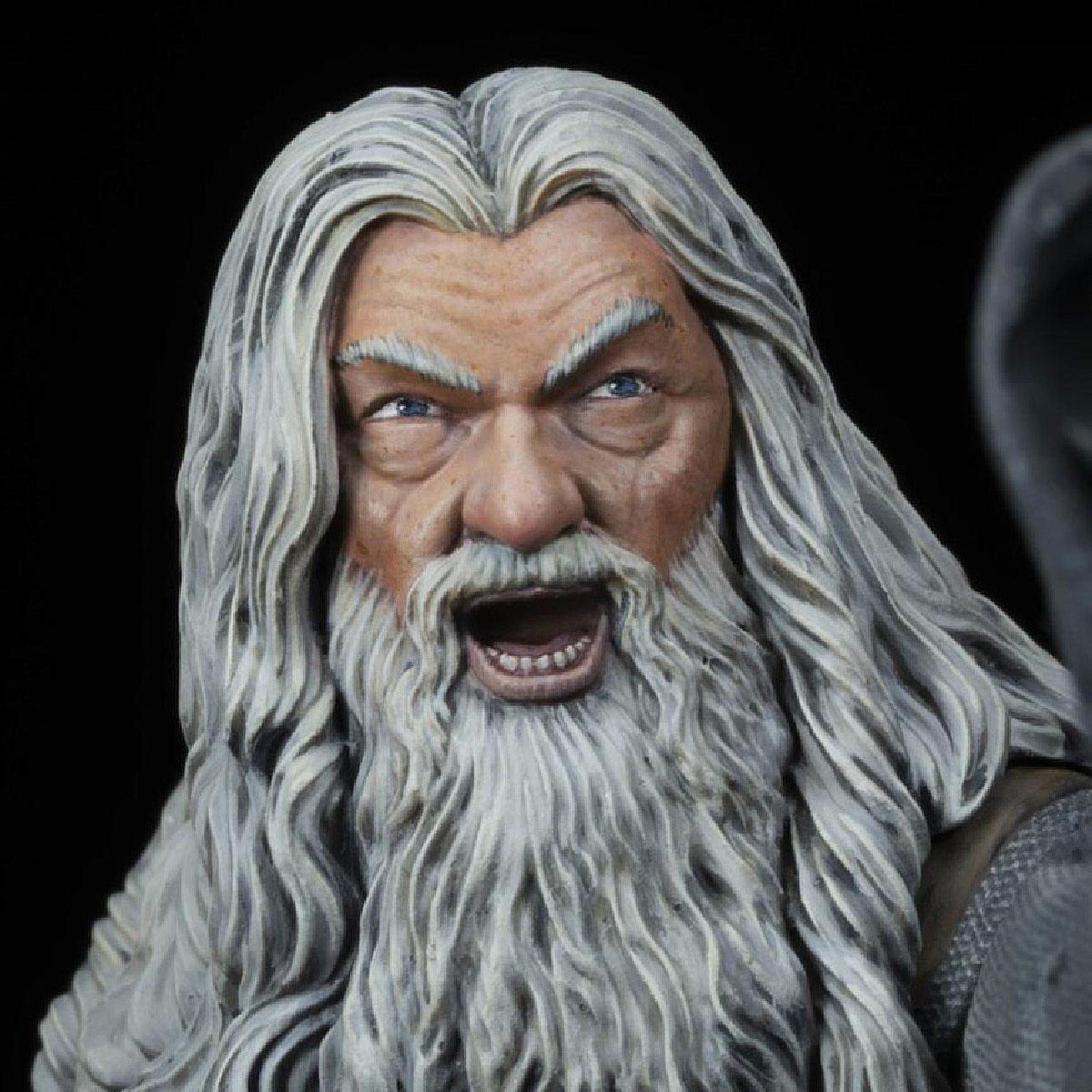 Primer plano de la cara de Gandalf. Barba blanca, pelo gris, boca abierta y ojos azules. Fondo oscuro.