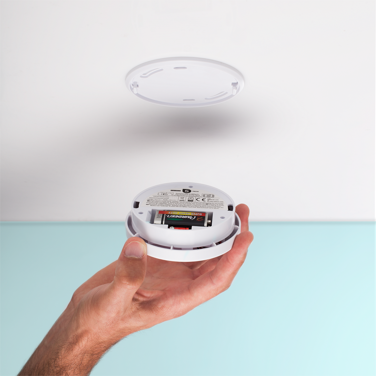 Una mano che tiene un rilevatore di fumo con batterie visibili. Sopra c'è un soffitto bianco.