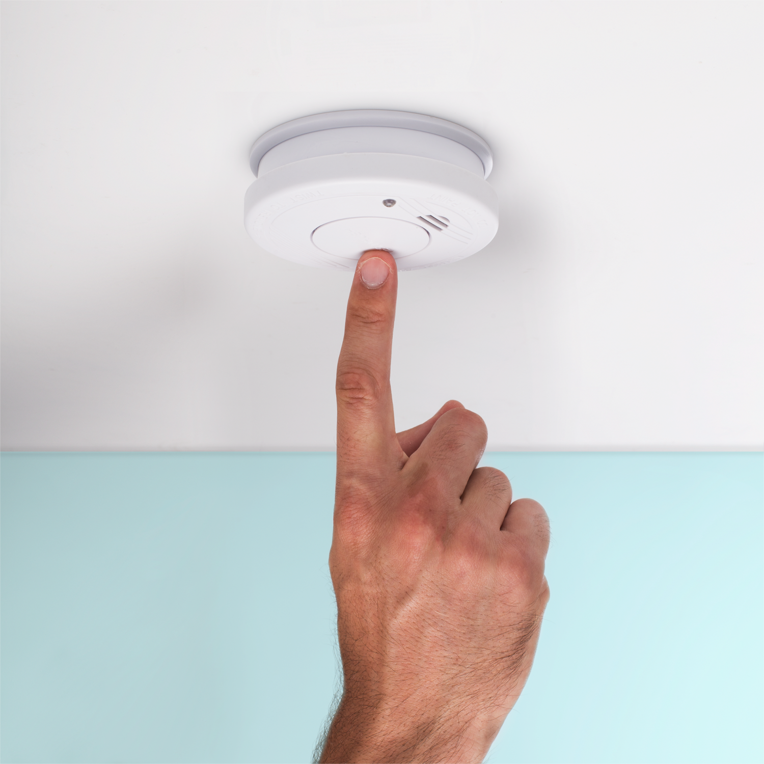 Un dito che preme il pulsante di un rilevatore di fumo bianco attaccato a un soffitto bianco.