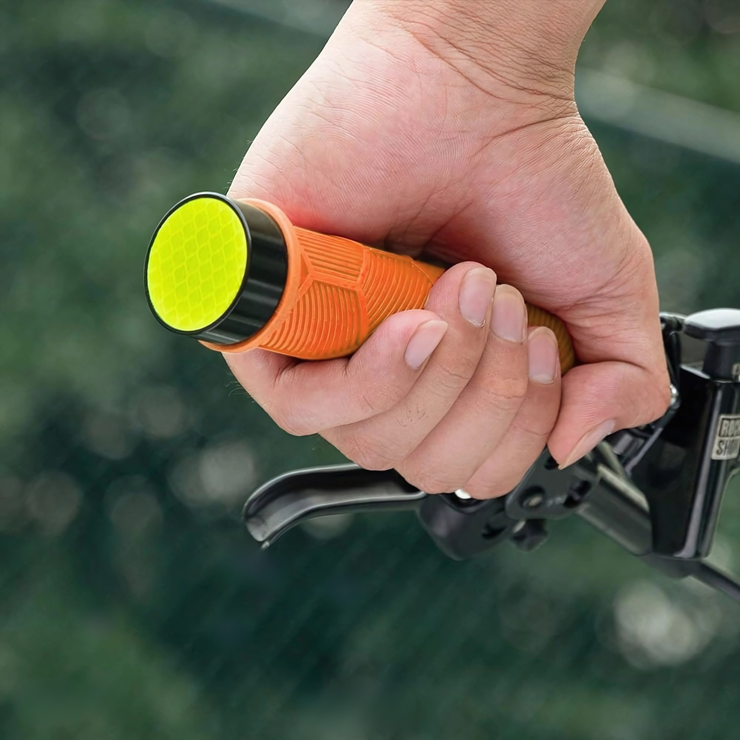 Dłoń trzyma pomarańczowy uchwyt rowerowy z żółtym odblaskiem. Widoczne części roweru.