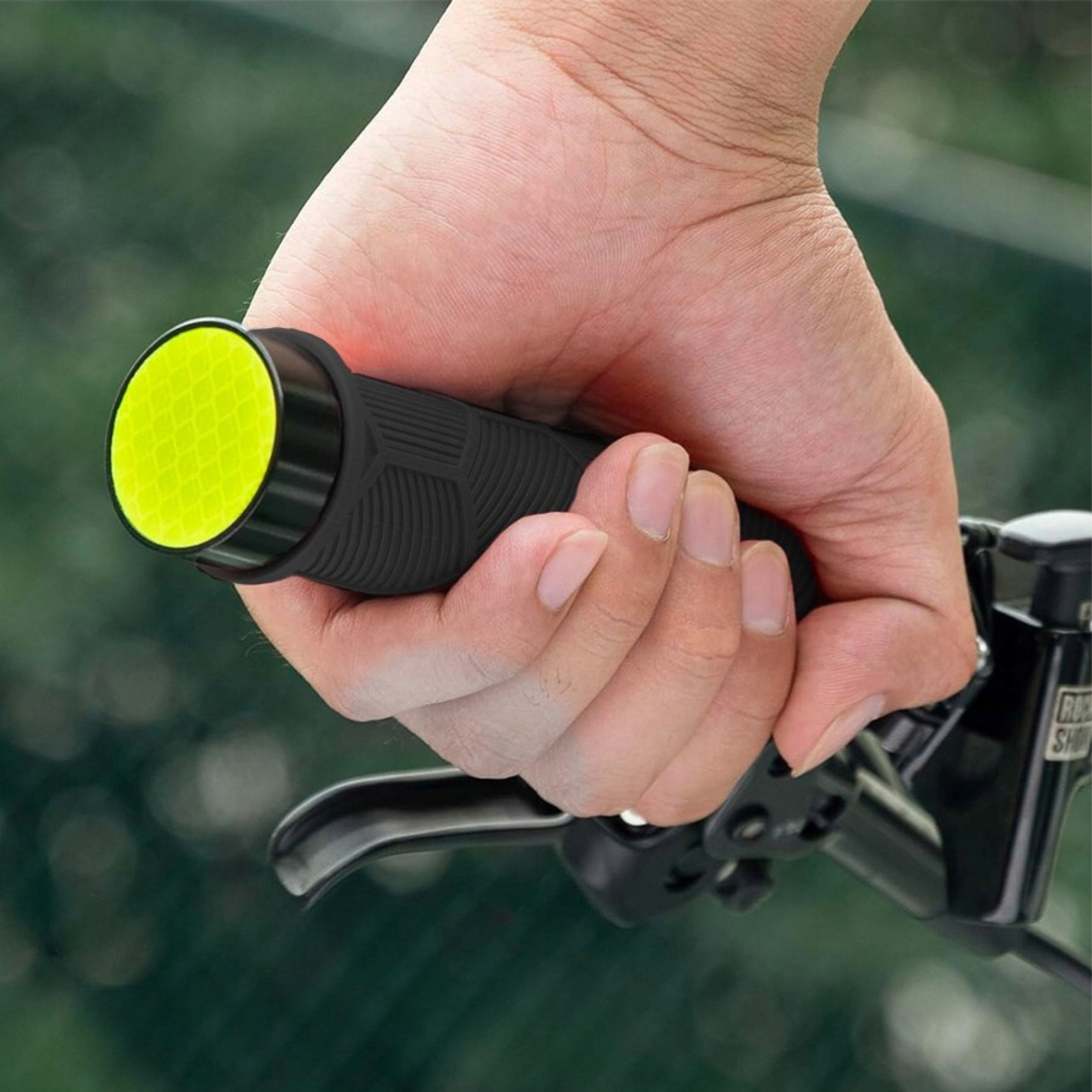 Dłoń trzyma czarny uchwyt rowerowy z żółtym odblaskiem. Widoczne części roweru.