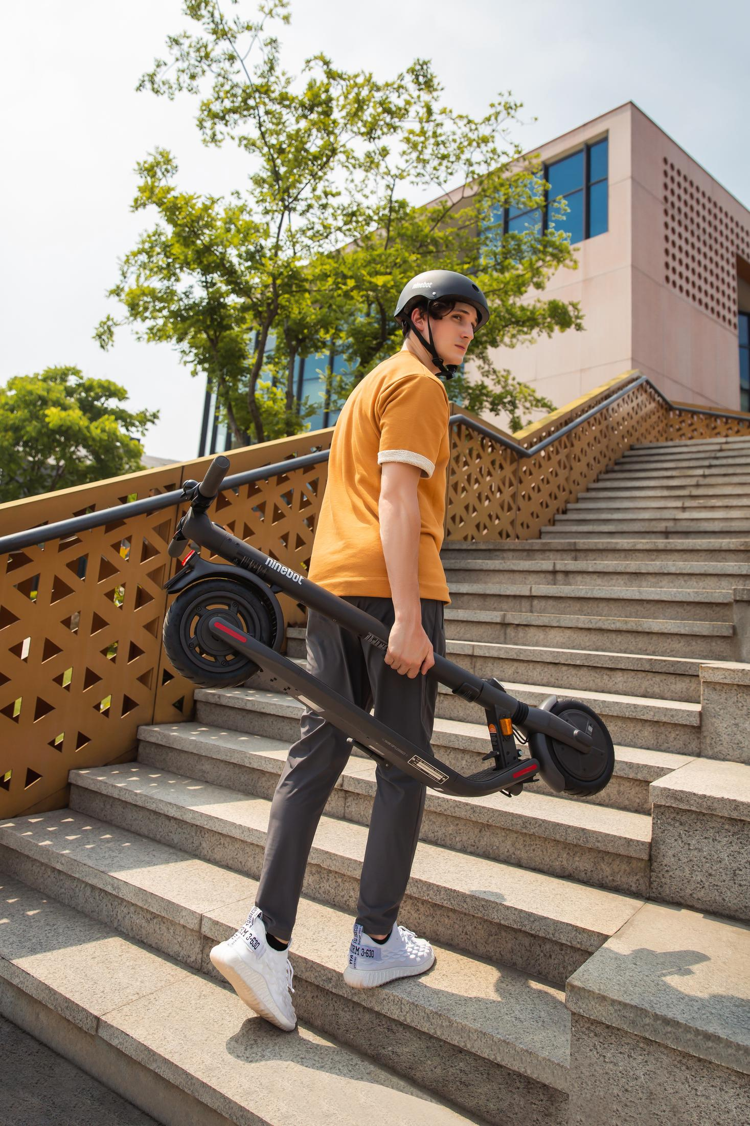 Mann trägt gefalteten Roller die Treppe hoch. Er trägt einen Helm und einen schwarzen Roller.