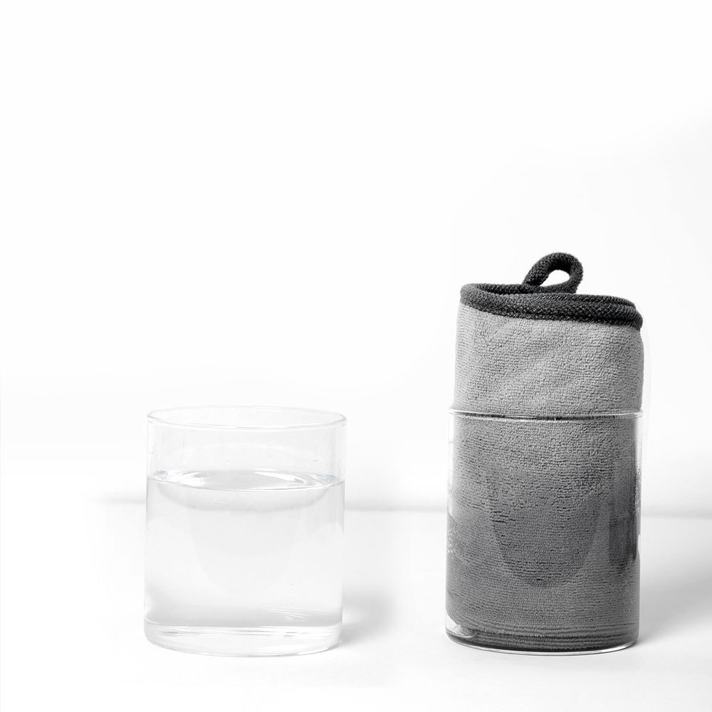 Szary ręcznik w przezroczystym szklanym pojemniku, obok szklanki z przezroczystym płynem.