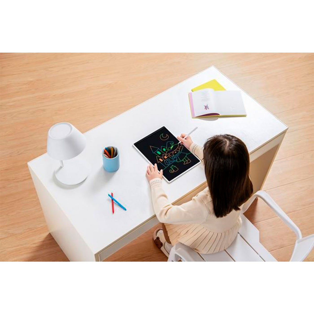 Dziecko rysuje na tablecie cyfrowym na białym stole. Jest lampa i ołówki.