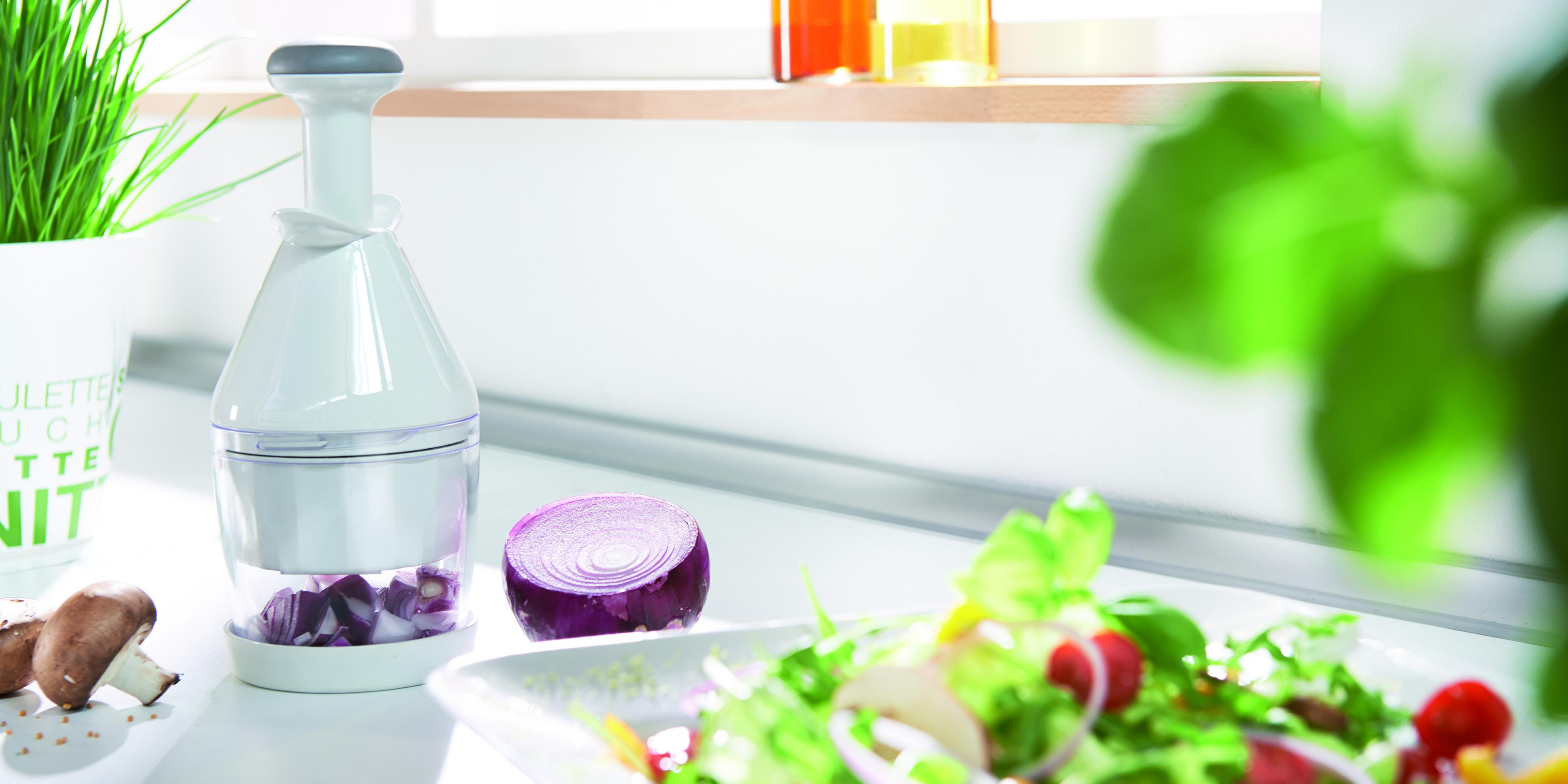 Ein weißer Zwiebelhacker neben einem Salat mit Fensterhintergrund, einer roten Zwiebelscheibe und Pilzen.