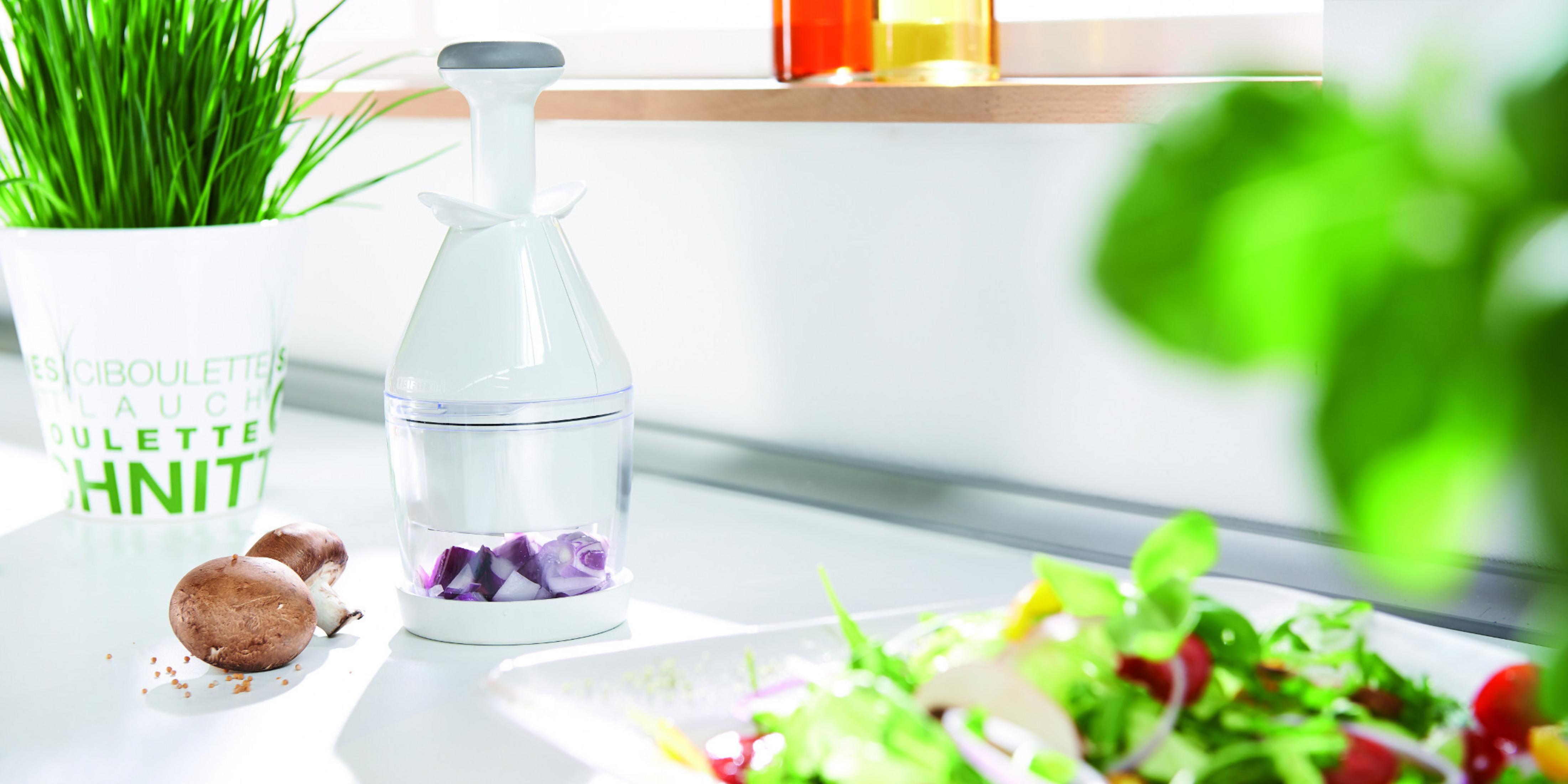 Ein weißer Zwiebelhacker neben einem Salat mit Fensterhintergrund, einer roten Zwiebelscheibe und Pilzen.