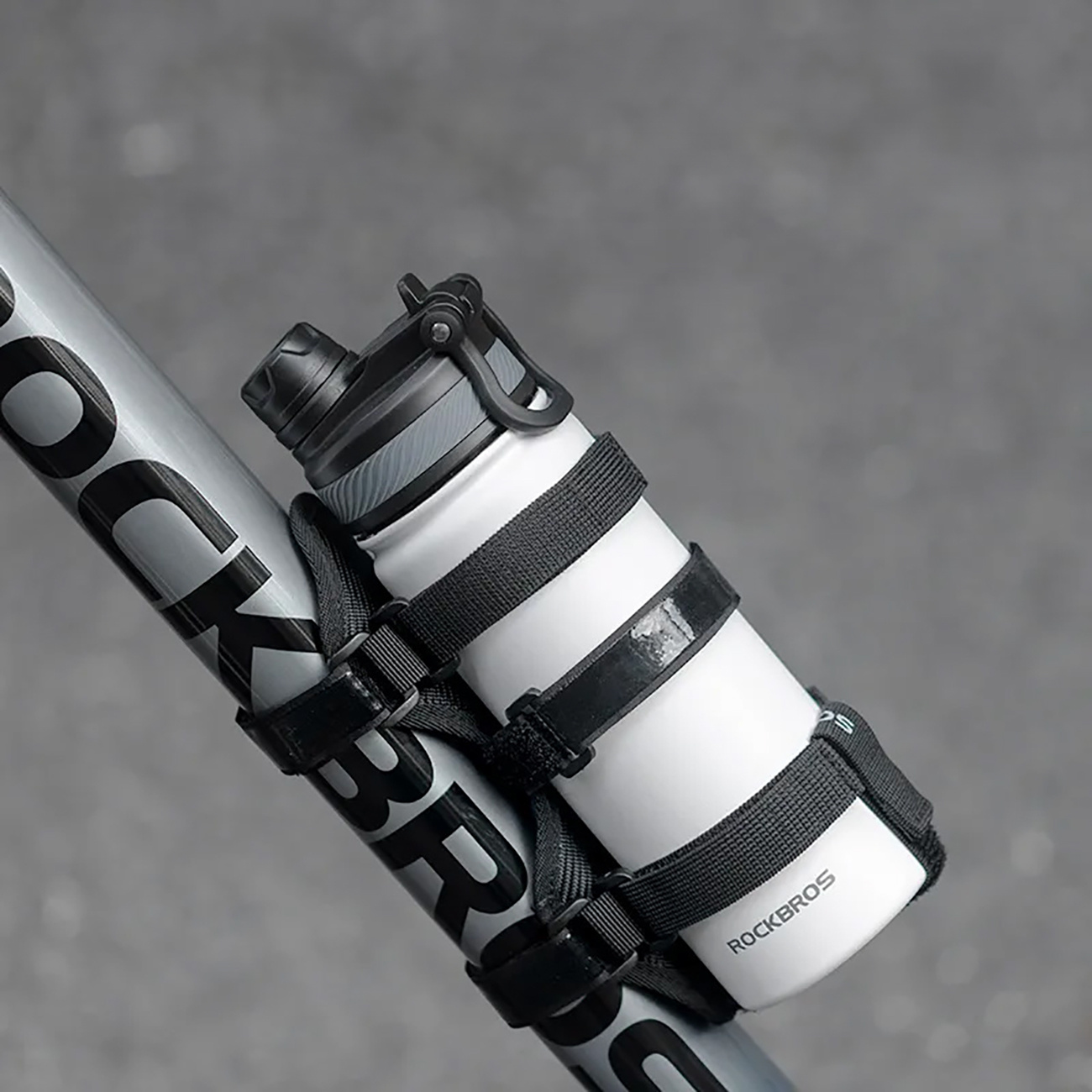 Biała butelka na wodę przymocowana do ramy roweru czarnymi paskami, widoczna nazwa marki.