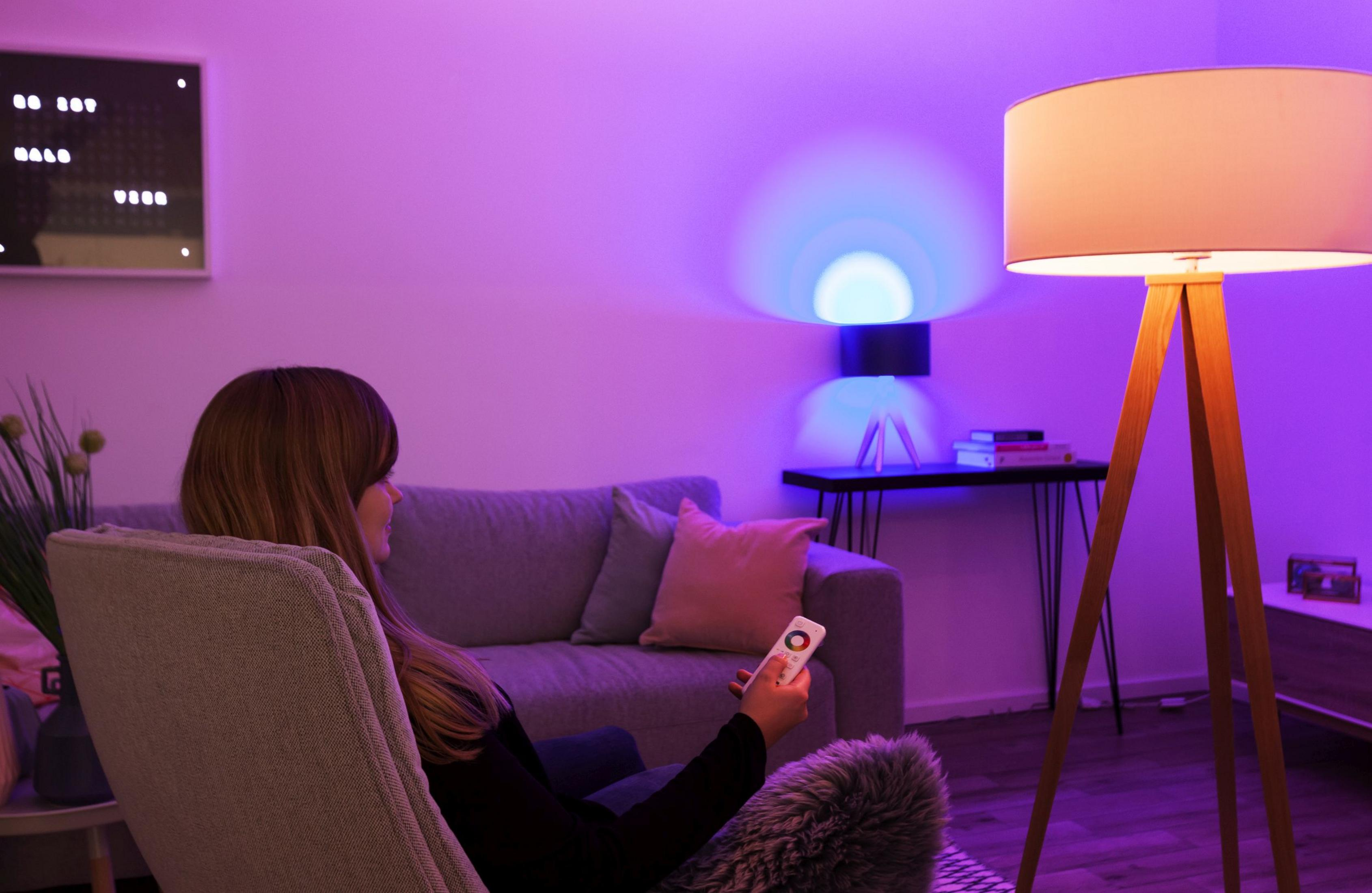 Frau sitzt mit Fernbedienung. Lampe rechts. Buntes Umgebungslicht.