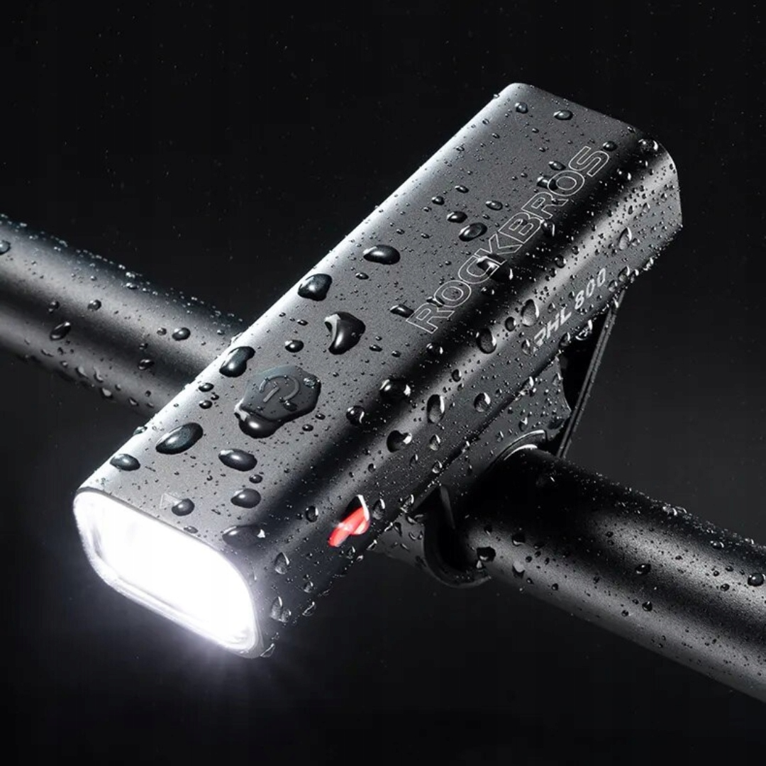 Czarna lampka rowerowa na kierownicy, z kroplami wody. Światło jest włączone. Widoczny jest napis "ROCKBROS RHL 800".