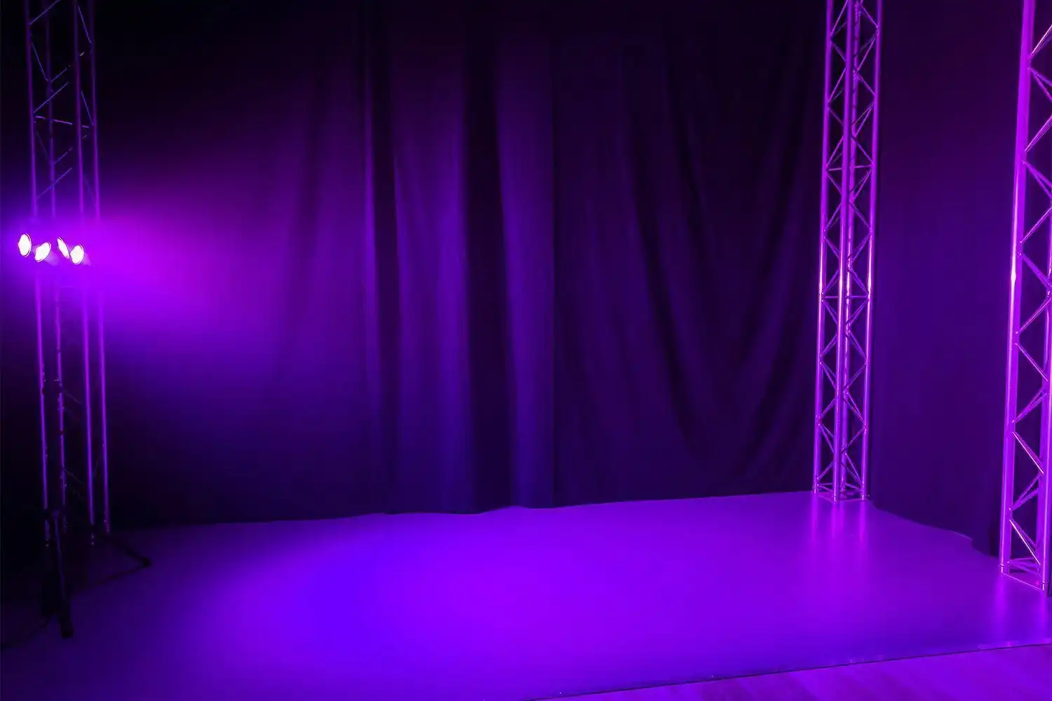 Un escenario iluminado con luz púrpura. Hay soportes metálicos a los lados. Un telón negro al fondo.