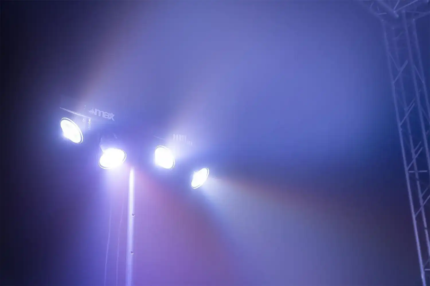 Cuatro luces en un poste emiten luz blanca brillante. Se ve el logo 'max'. El fondo es azul brumoso.