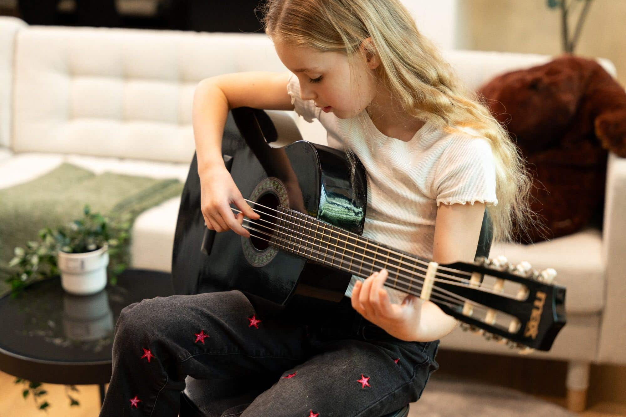Niña tocando guitarra negra, con jeans de estrellas, sentada en un sofá.