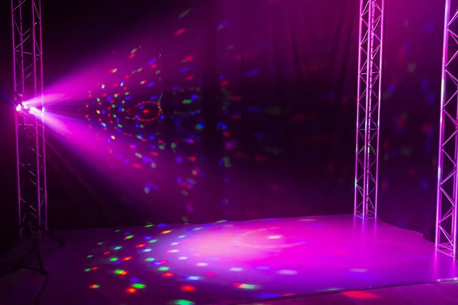 Un escenario iluminado con luz rosa. Dos armazones de metal enmarcan el escenario. Luces de colores se proyectan.