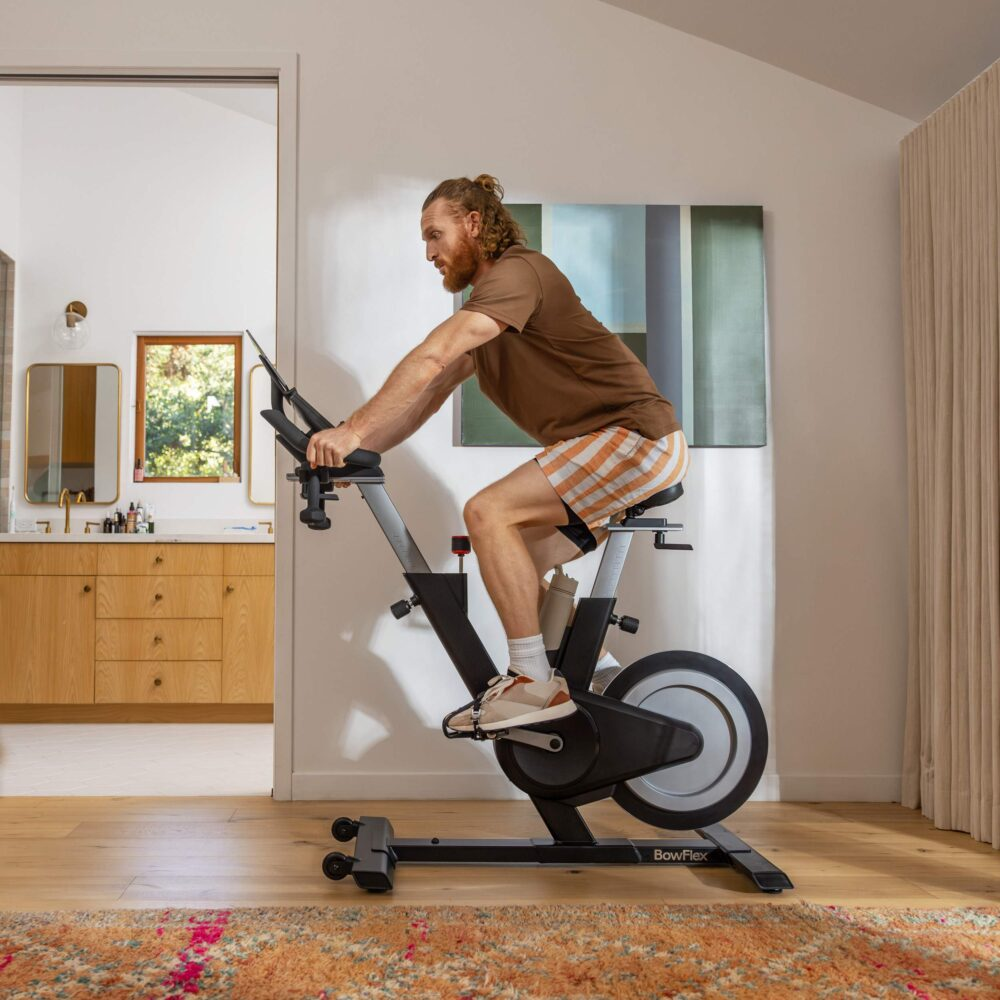 Un hombre haciendo ejercicio en una bicicleta estática en casa, con una alfombra y un espejo.