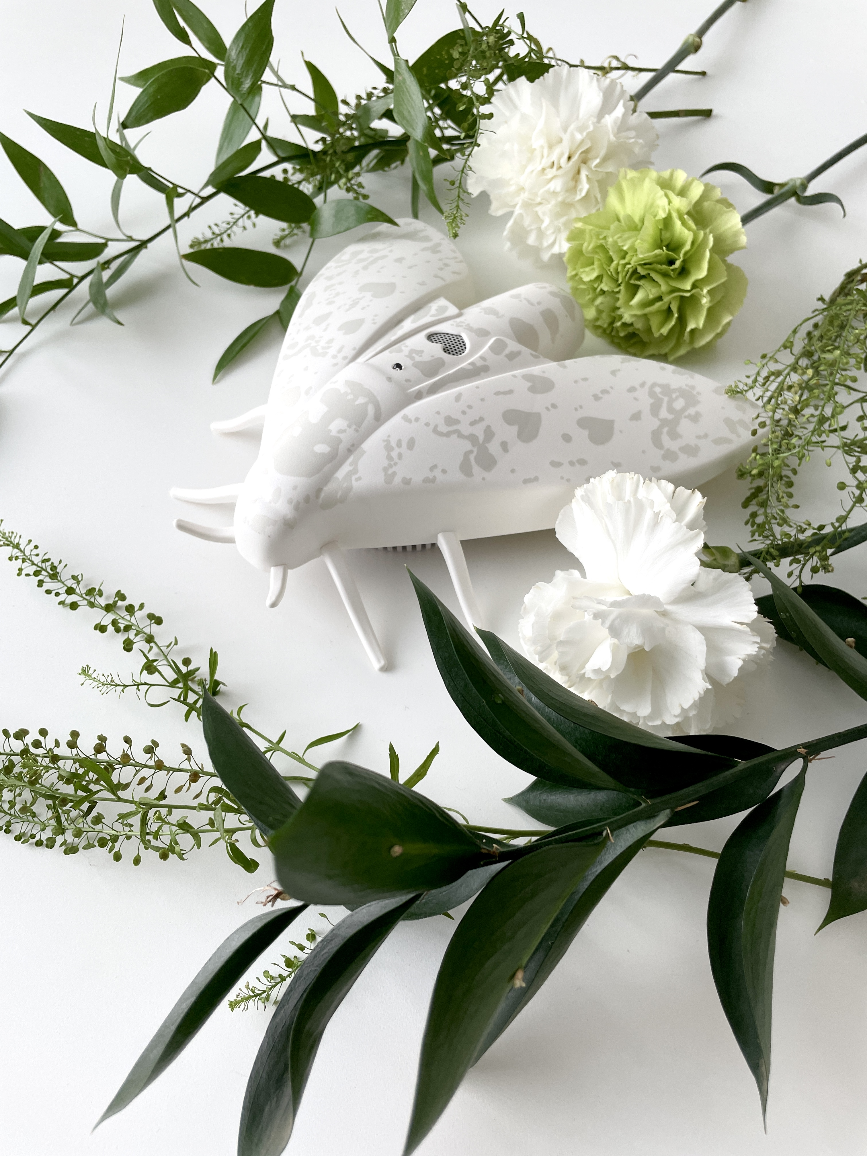 Eine weiße, abstrakte Insektenskulptur, umgeben von weißen Blumen und grünem Laub, auf weißem Hintergrund.
