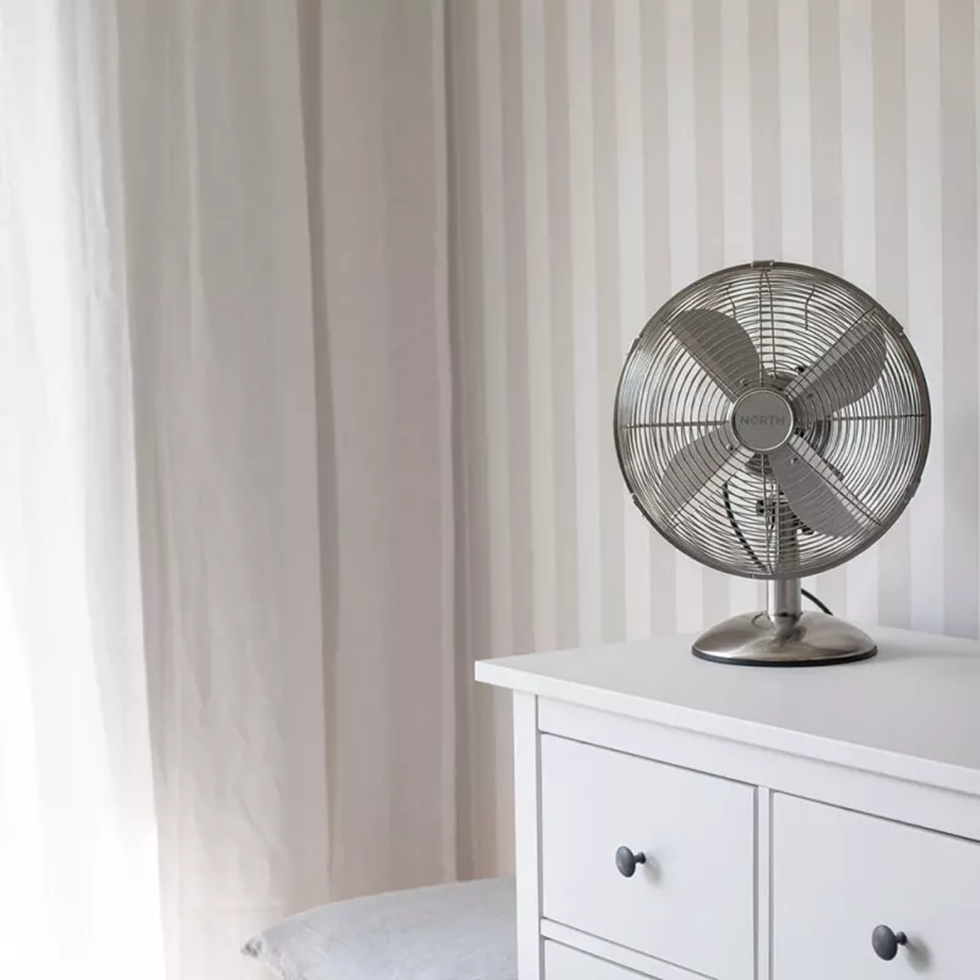 Ventilatore argentato su una cassettiera bianca. Tende chiare a sinistra, carta da parati a strisce sullo sfondo.