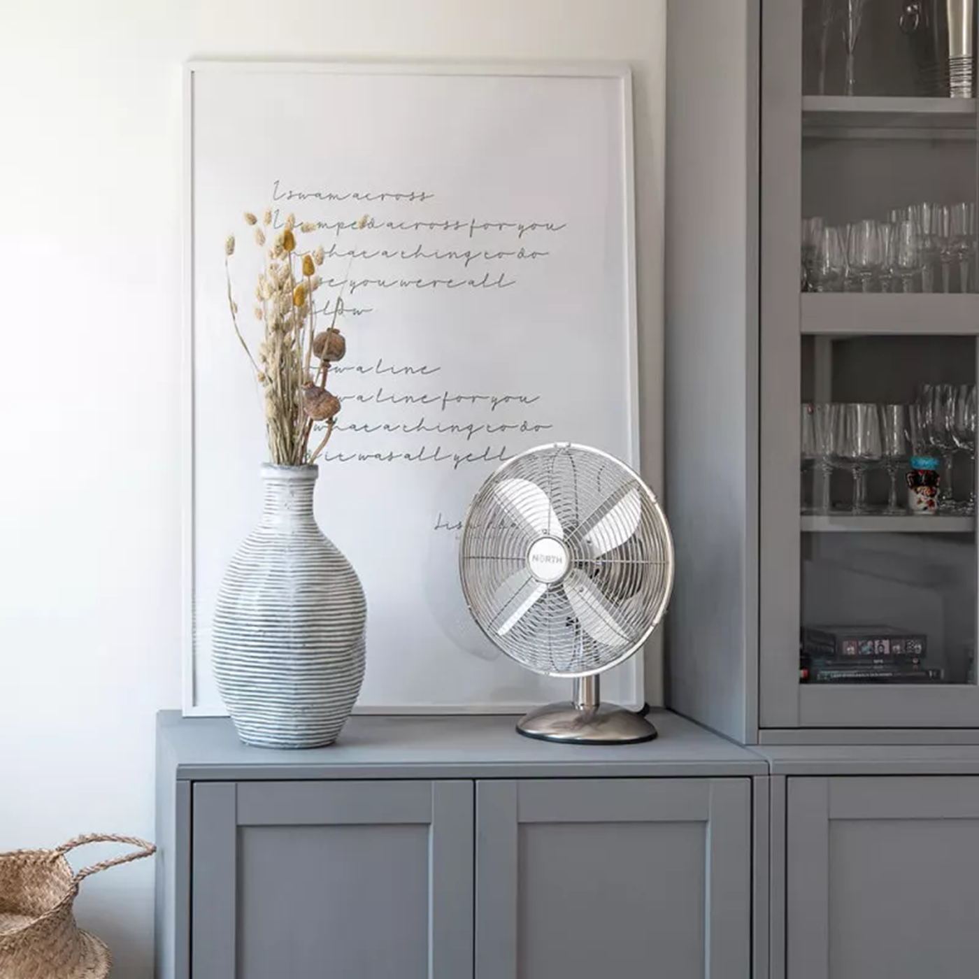 Ventilatore argentato su mobile con vaso e arte incorniciata. Vetrina a destra.