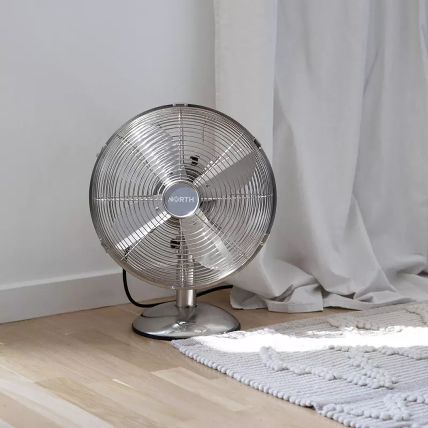 Ventilatore argentato su pavimento in legno. Un tappeto bianco e una tenda chiara sullo sfondo.