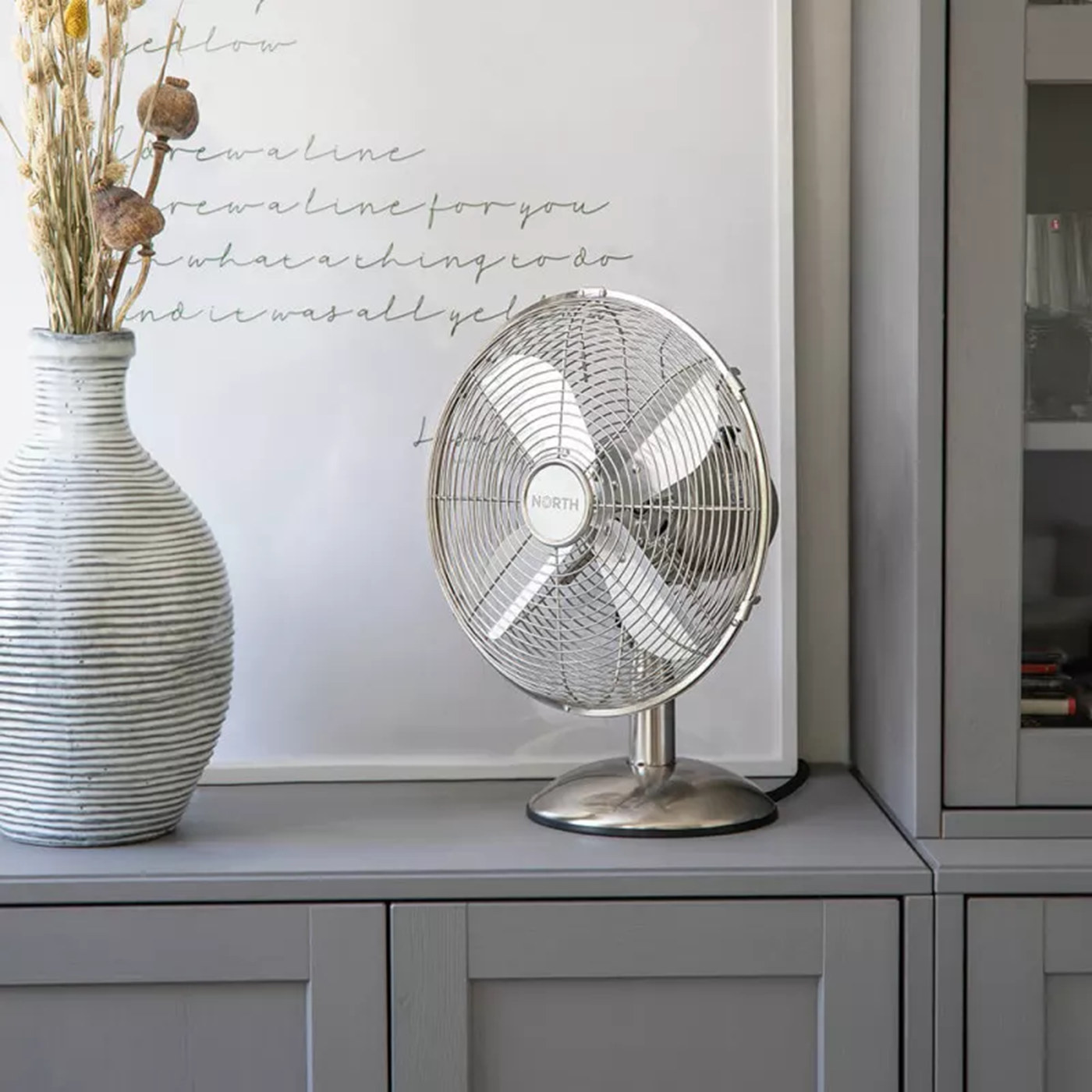 Ventilatore argentato su mobile con vaso e arte incorniciata. Vetrina a destra.