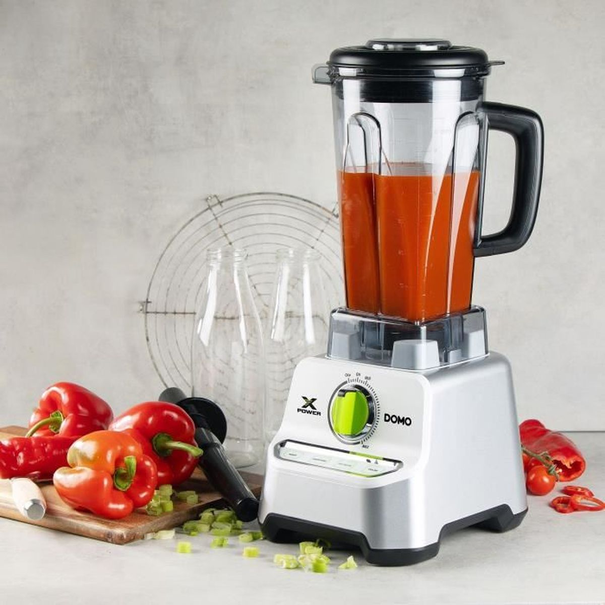 Un frullatore con liquido rosso all'interno, circondato da verdure e bottiglie trasparenti.