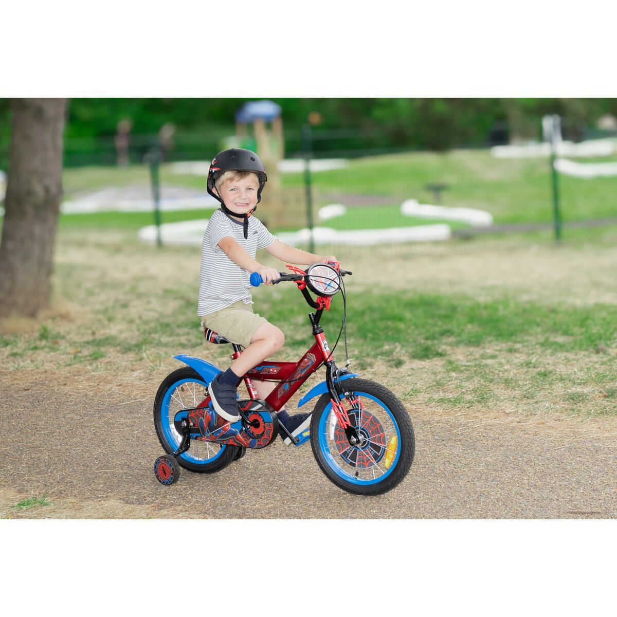 Un bambino guida una bici rossa di Spiderman su un sentiero di ghiaia. Indossa un casco. Lo sfondo è un parco verde.