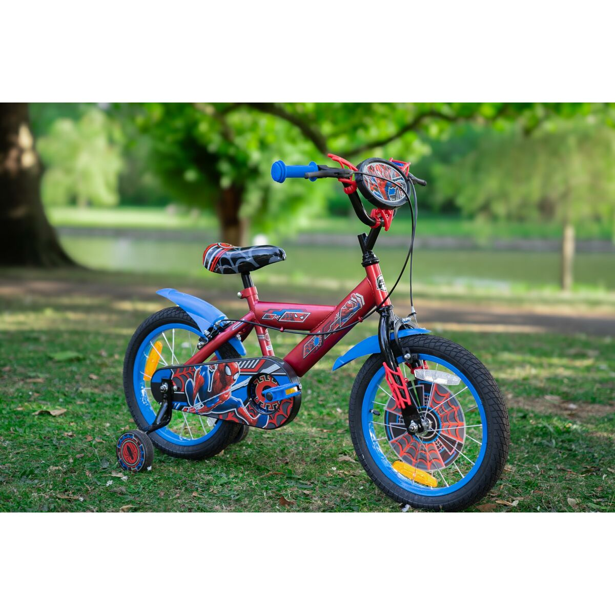 Una bici rossa di Spiderman sull'erba. Lo sfondo presenta alberi e un lago. La bici ha dettagli blu.