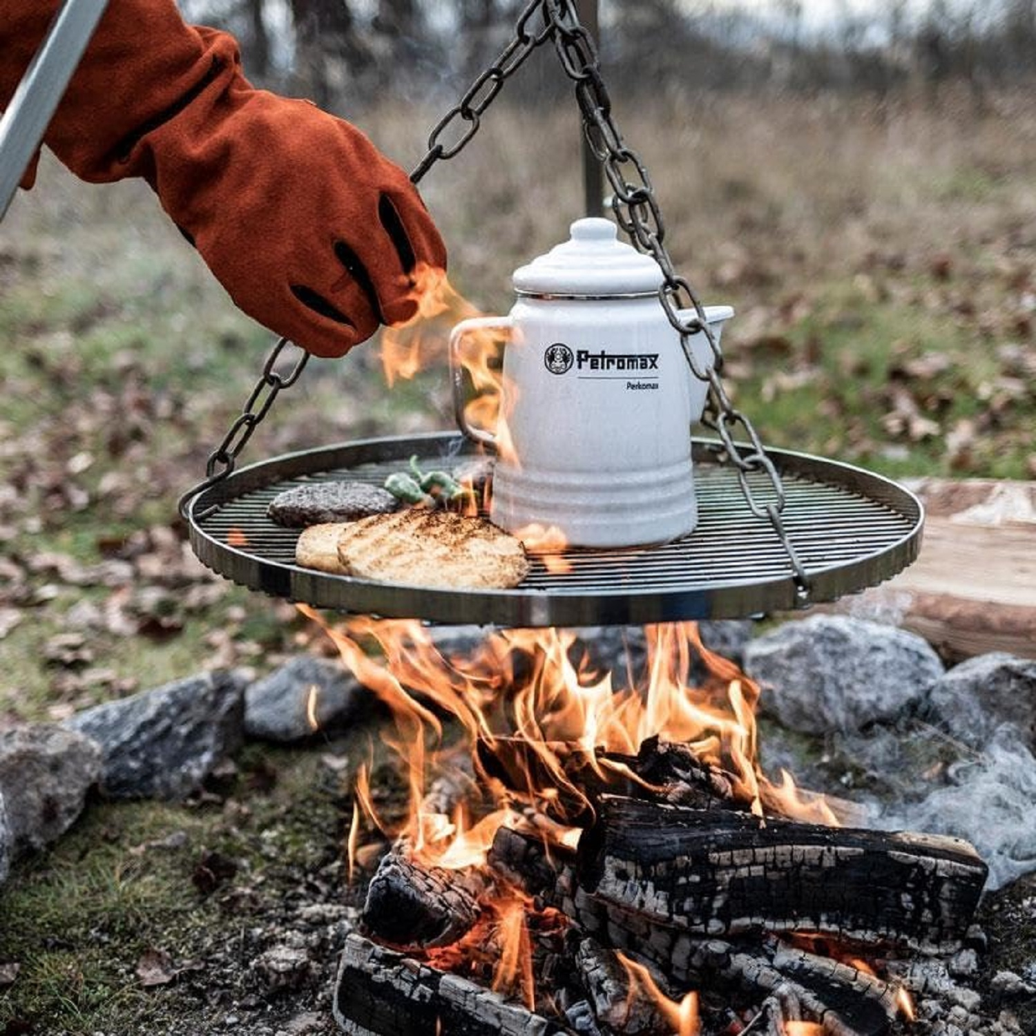 Ein Grill über einem Feuer mit Essen und einem Kessel. Eine behandschuhte Hand greift nach dem Kessel. Der Hintergrund ist natürlich.