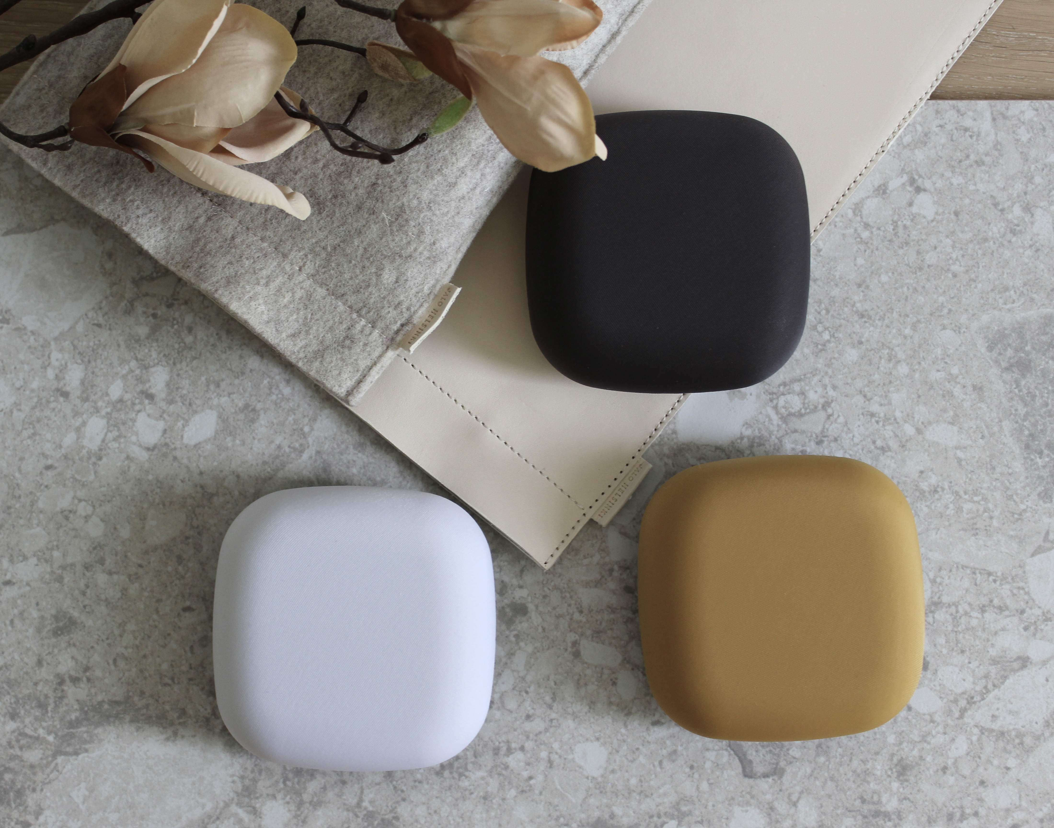 Drei farbige, quadratische, berührungsempfindliche Pads auf einer grauen Oberfläche, mit einem beige, gefalteten Tuch und Blumen.