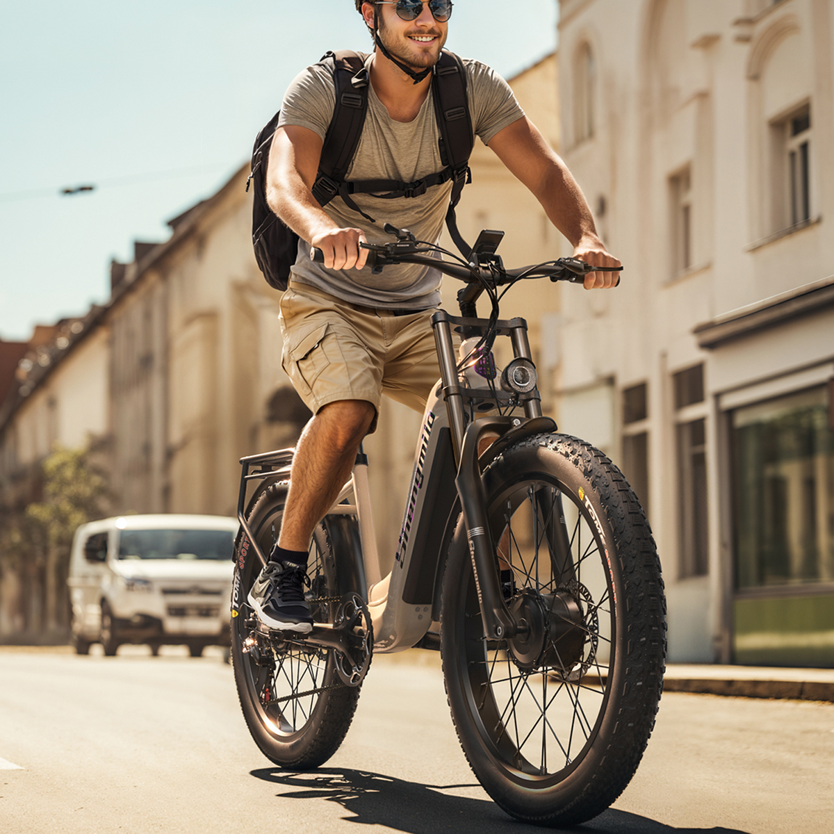 Mężczyzna jedzie na beżowym rowerze elektrycznym ulicą. Nosi kask, okulary przeciwsłoneczne i plecak. Budynki w tle.