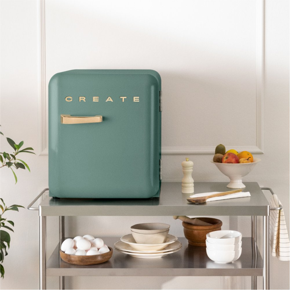Una nevera color verde azulado con un mango de madera y la palabra CREATE en un carrito de metal con frutas y huevos.