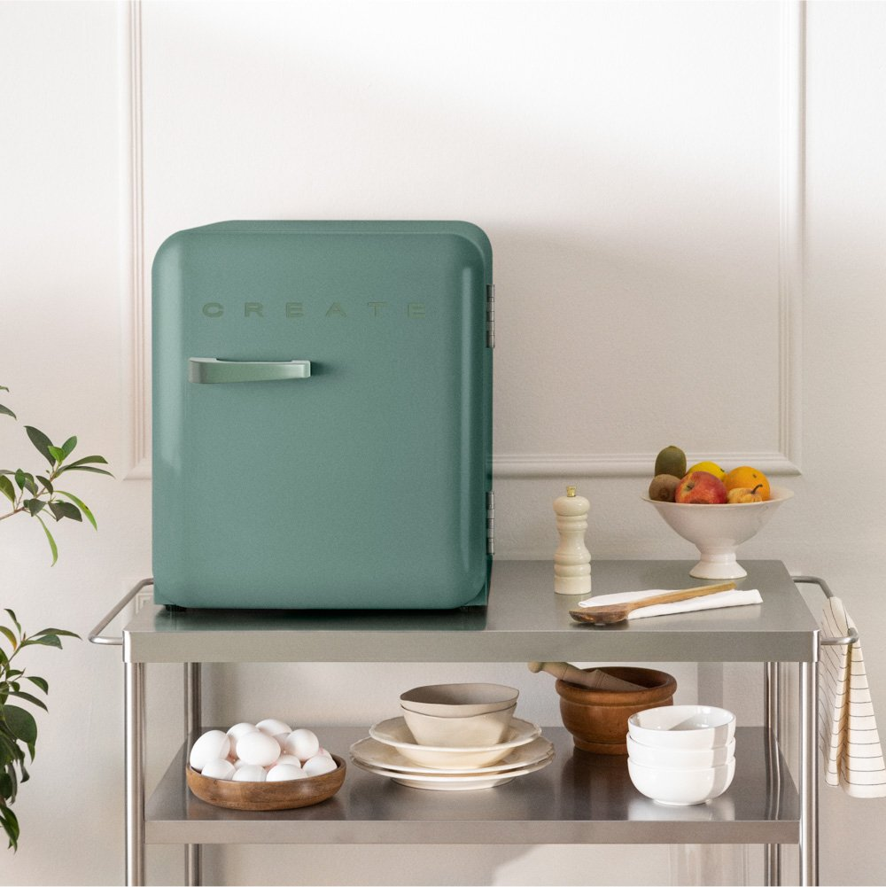 Una nevera color verde azulado con un mango plateado y la palabra CREATE en un carrito de metal con frutas y huevos.