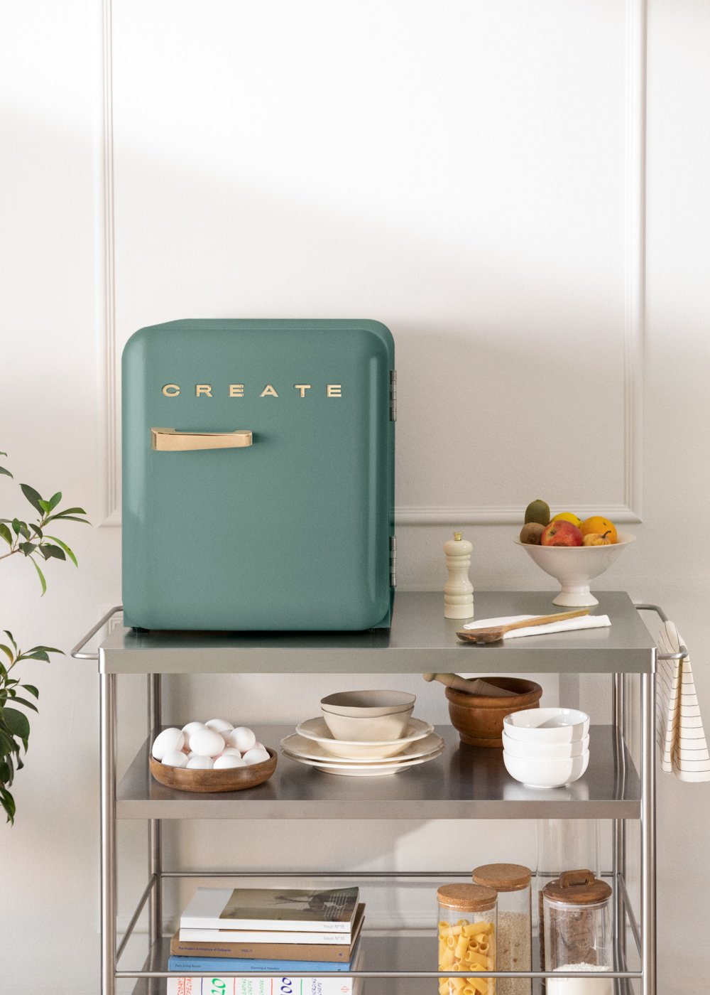 Una nevera color verde azulado con un mango de madera y la palabra CREATE en un carrito de metal con frutas, huevos y libros.