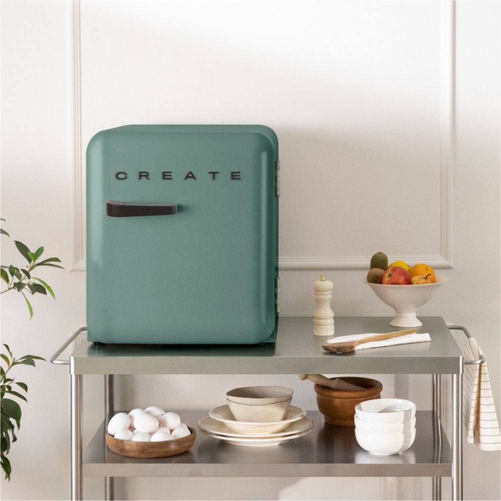 Una nevera color verde azulado con un mango negro y la palabra CREATE en un carrito de metal con frutas y huevos.