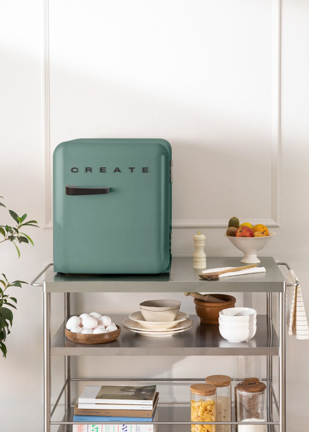 Una nevera color verde azulado con un mango negro y la palabra CREATE en un carrito de metal con frutas, huevos y libros.