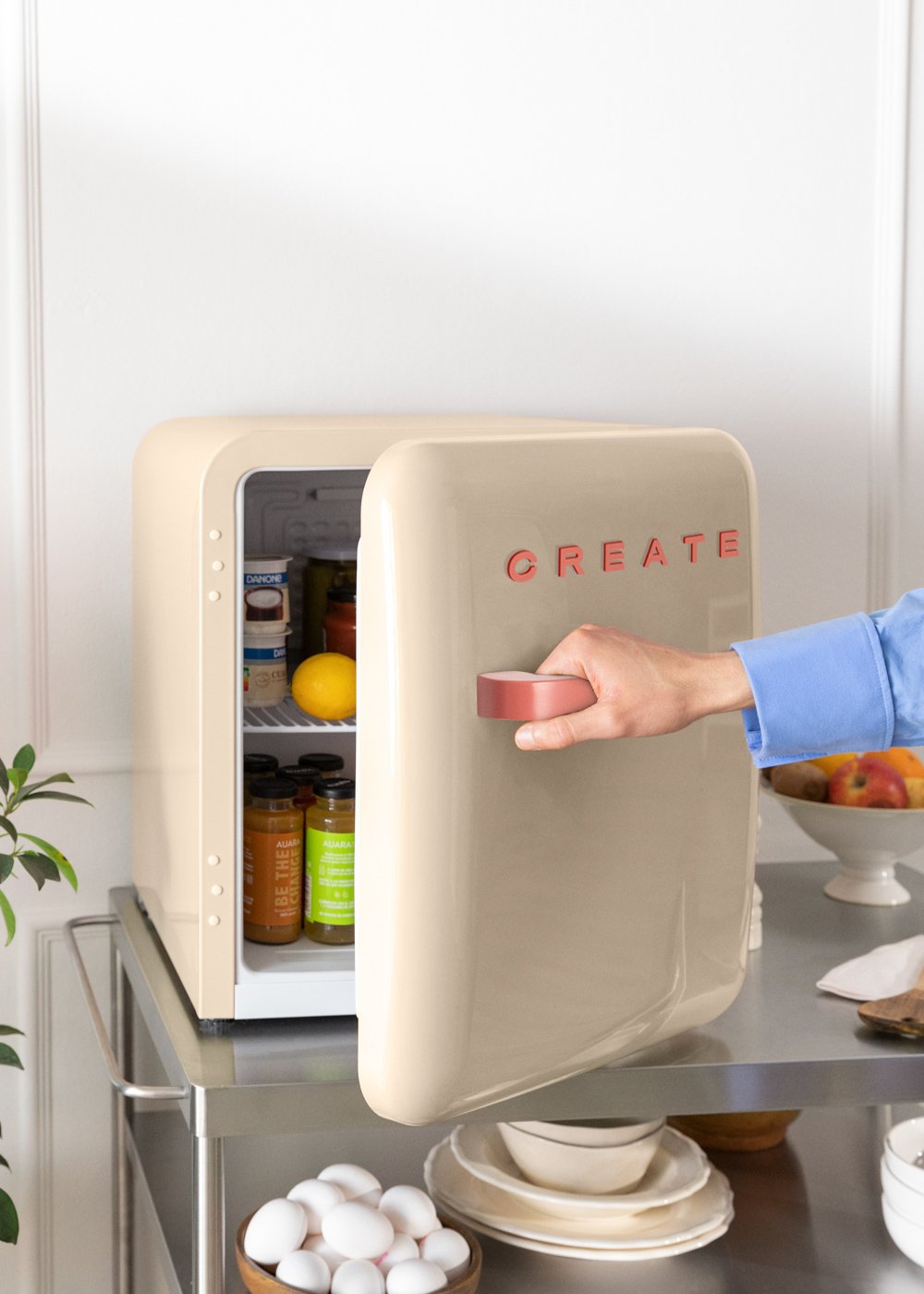 Un mini-refrigerador beige con 'CREATE' rojo abierto, revelando su contenido. Una mano abre la puerta.