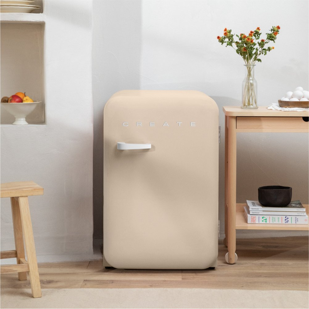 Un mini-refrigerador beige con 'CREATE'. Una mesa de madera, un taburete y un jarrón de flores a su lado.
