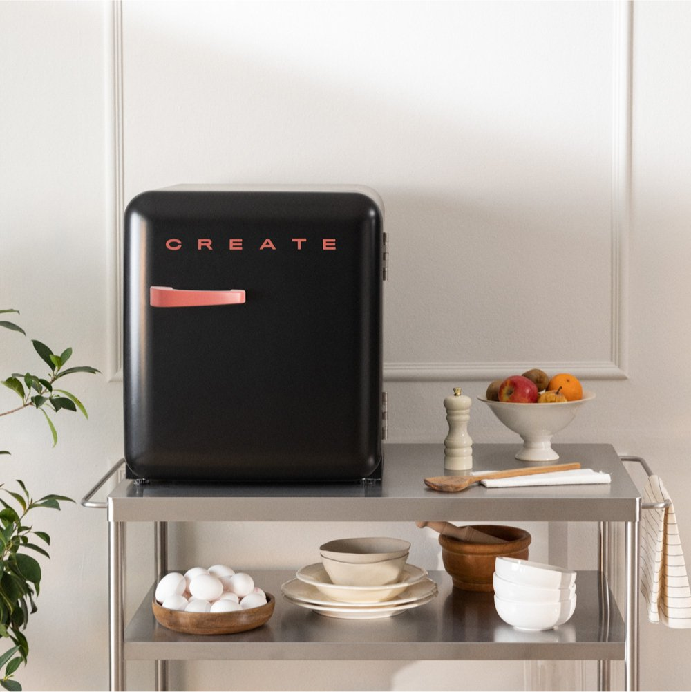 Un mini-refrigerador negro con 'CREATE' rosa sobre un carrito de acero inoxidable. Frutas, huevos y utensilios presentes.