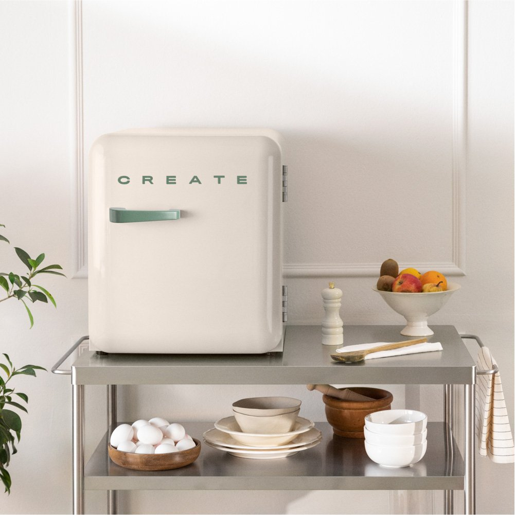 Un mini-refrigerador beige con 'CREATE' verde sobre un carrito de acero inoxidable. Frutas, huevos y utensilios presentes.