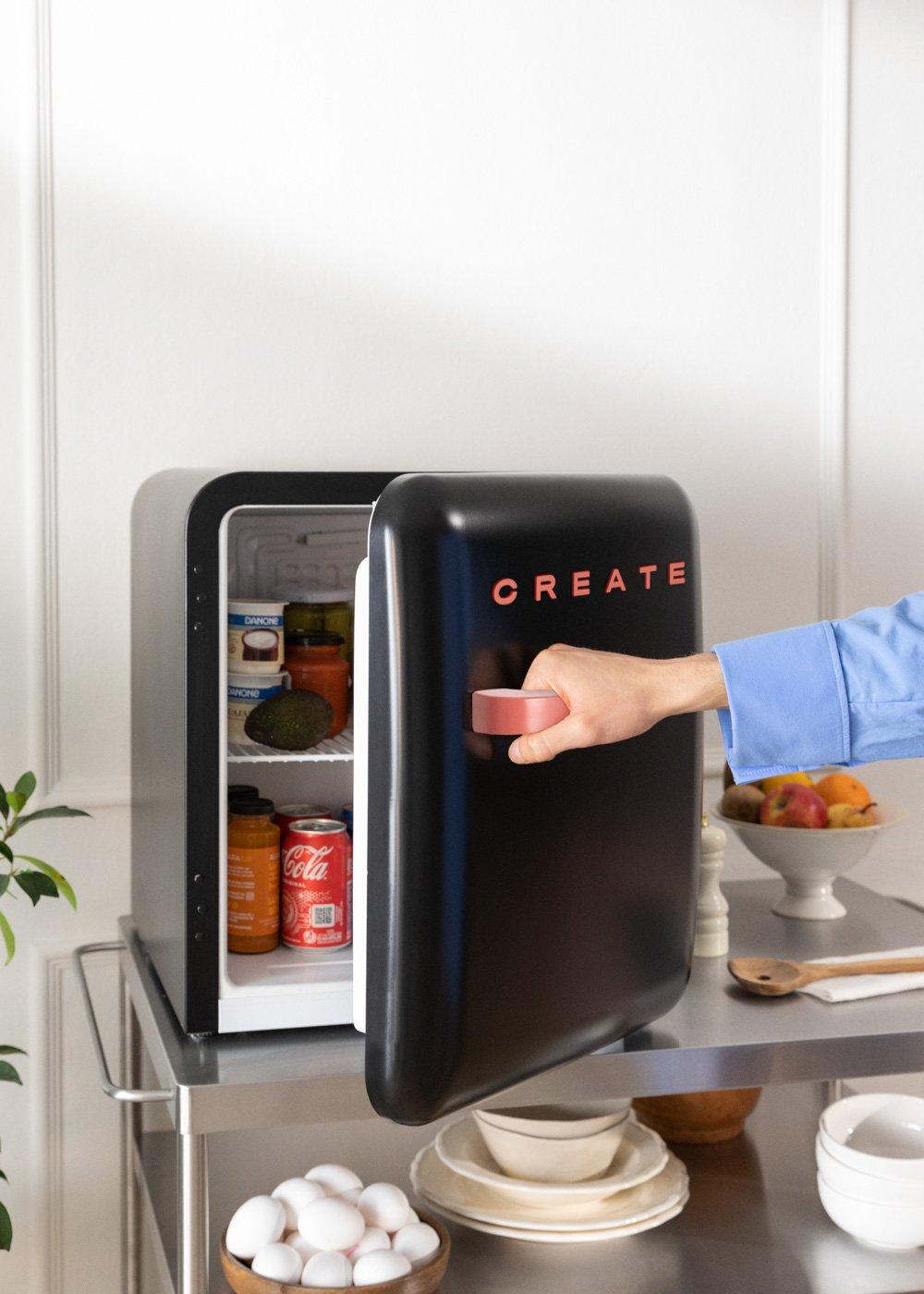 Un mini-refrigerador negro con 'CREATE' abierto. Una mano lo abre, revelando elementos dentro. Está en un carrito de acero.