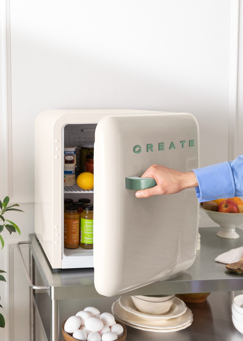 Un mini-refrigerador beige con 'CREATE' verde abierto. Una mano lo abre, revelando su contenido. En un carrito de acero.